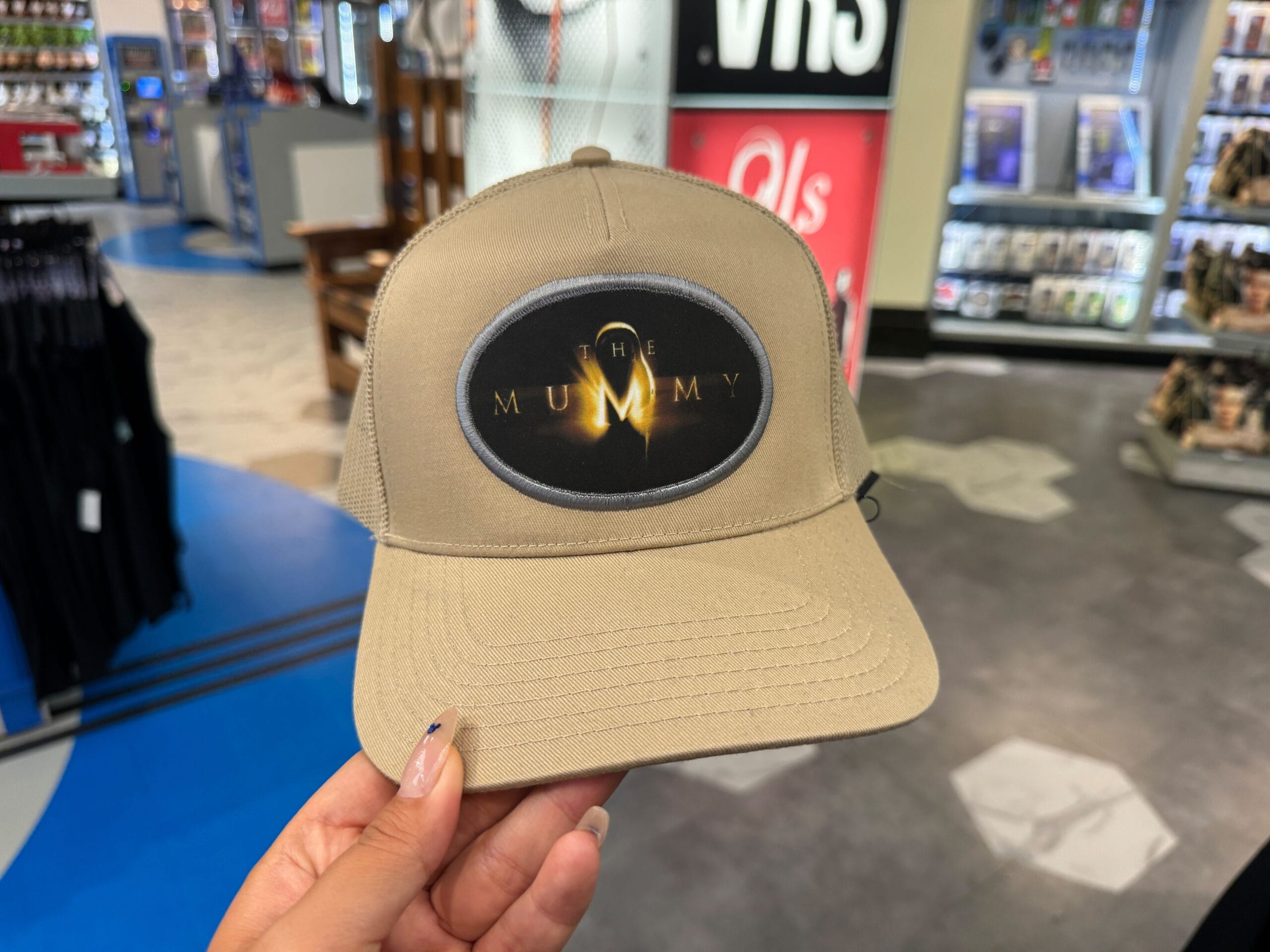 A hand holds a tan trucker hat from the UNIVRS Mummy Collection with "The Mummy" logo inside a retail store.