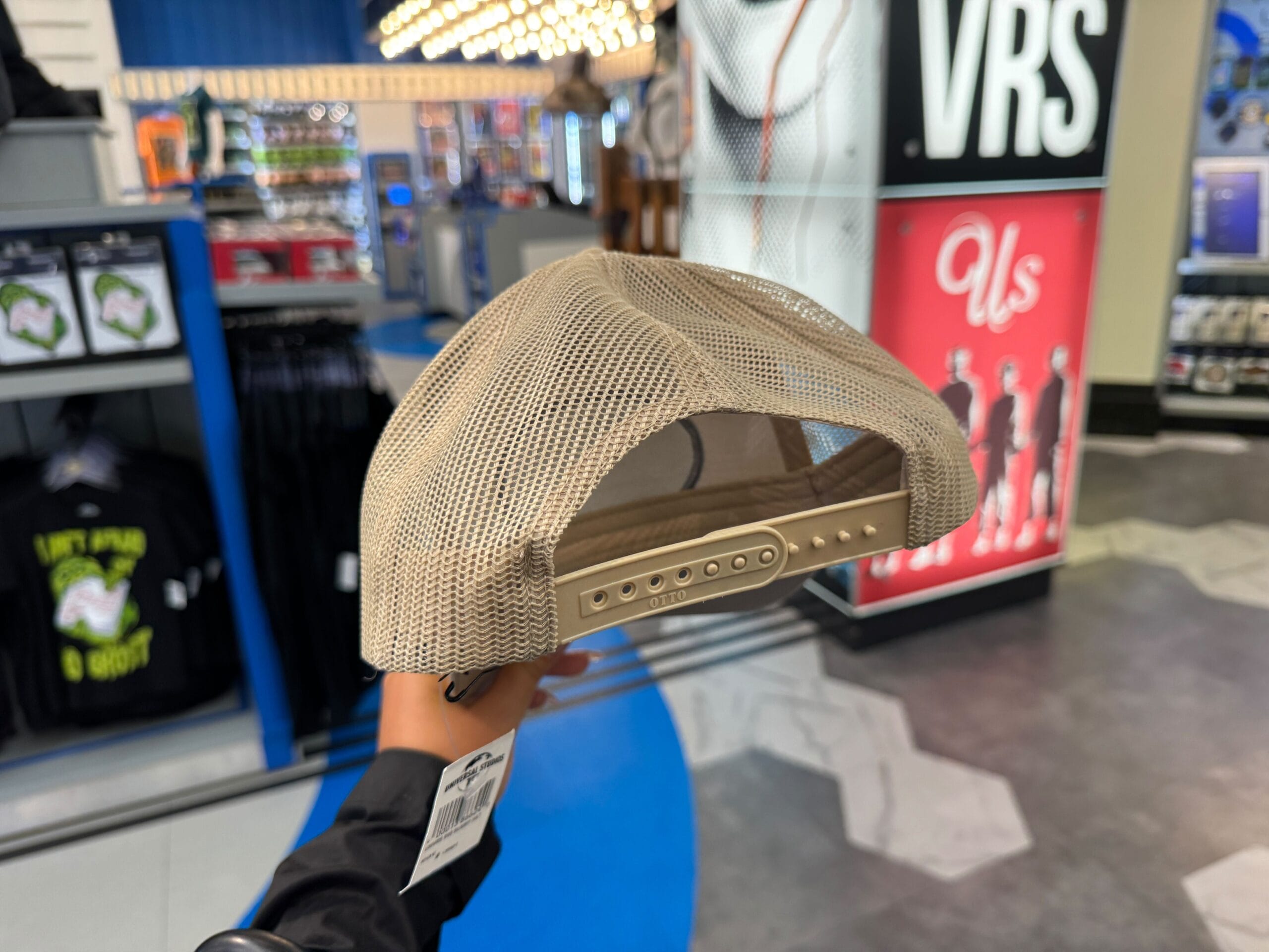 A hand holds a beige mesh snapback cap from the Mummy Collection in a UNIVRS store with clothing and signs behind.