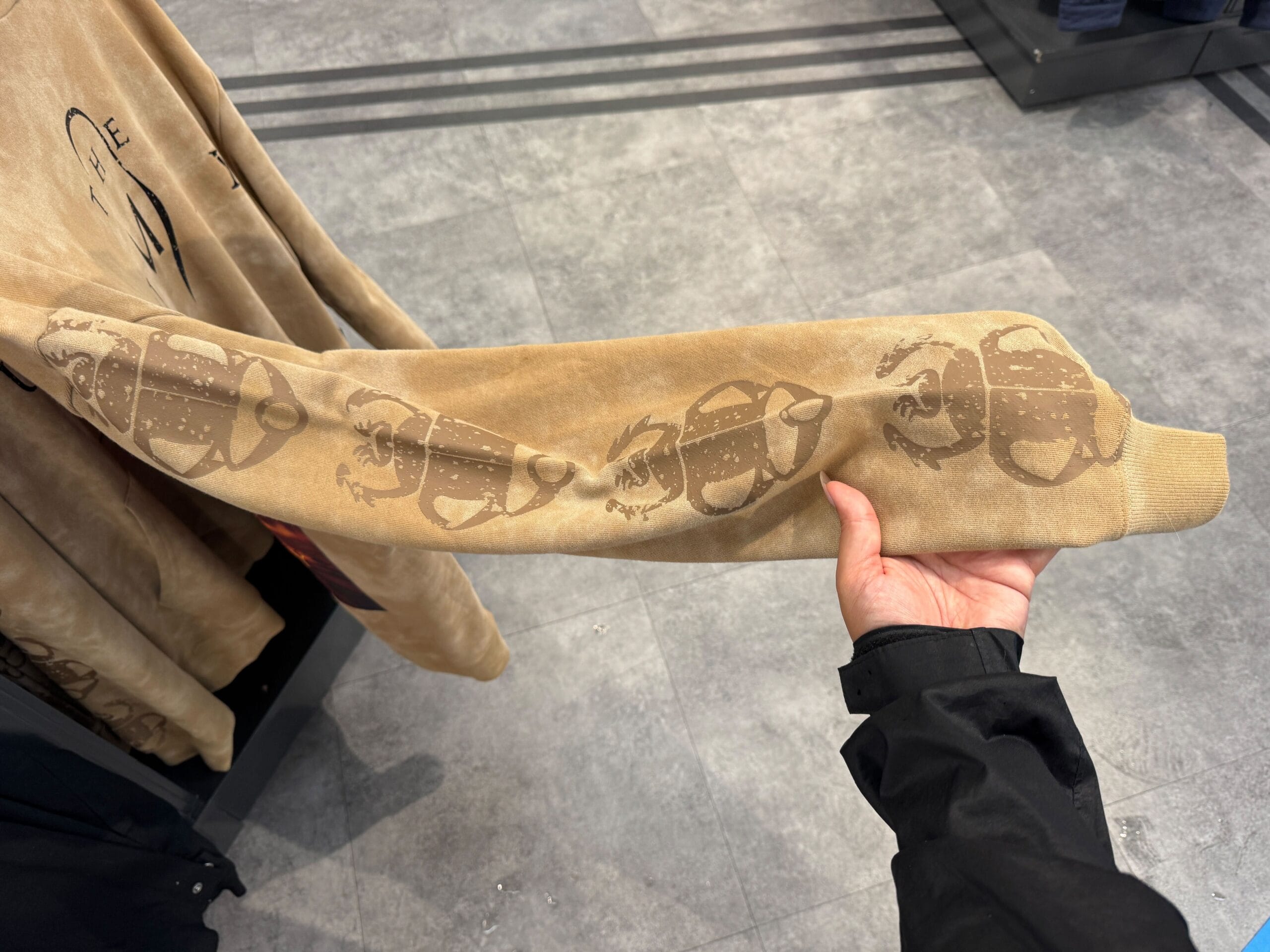 A hand holds a tan sweatshirt sleeve with a repeating beetle design from the UNIVRS Mummy Collection in a clothing store.