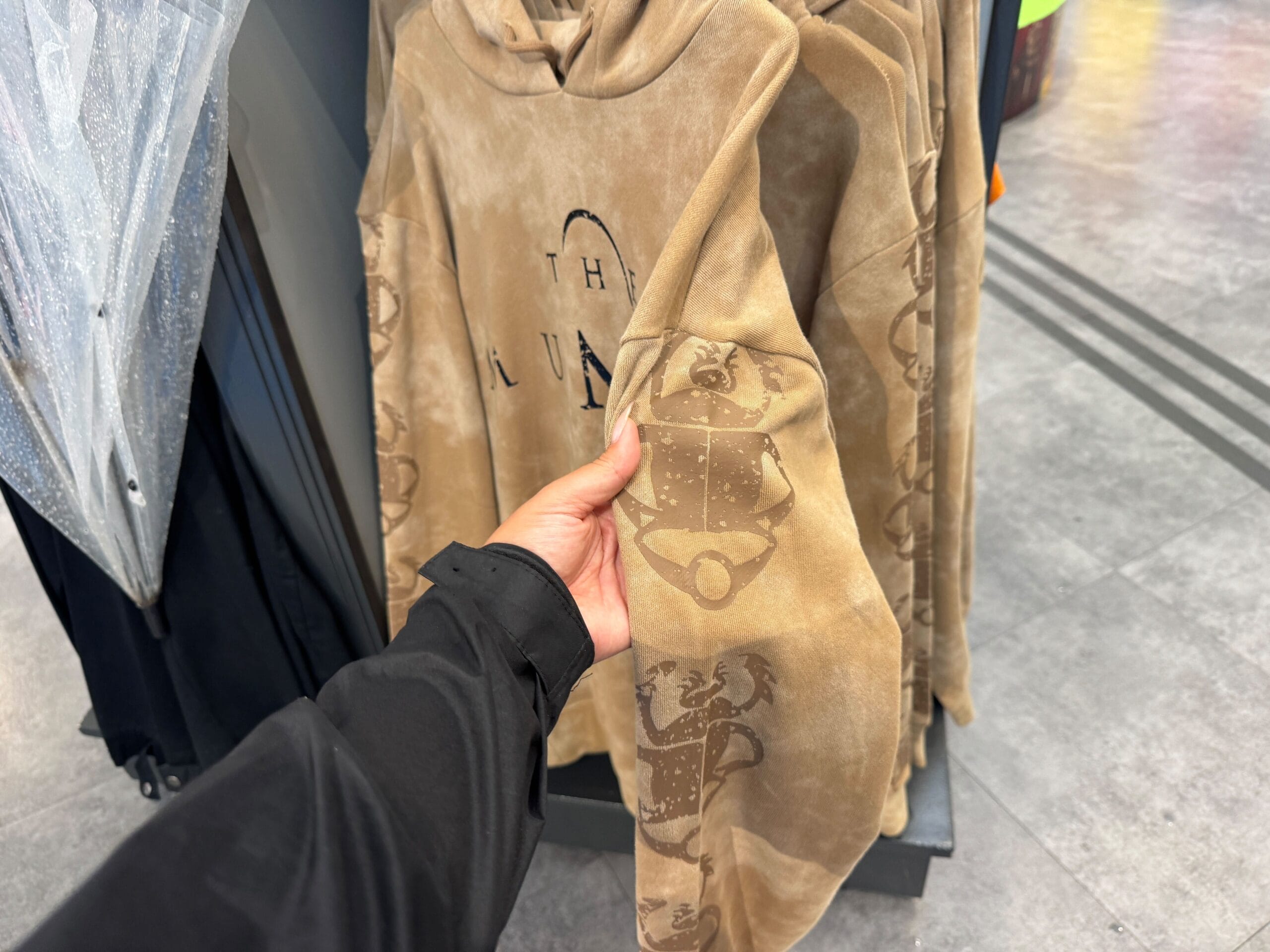 A hand holds a tan hoodie from the UNIVRS Mummy Collection with faded designs and sleeve patterns in a clothing store.