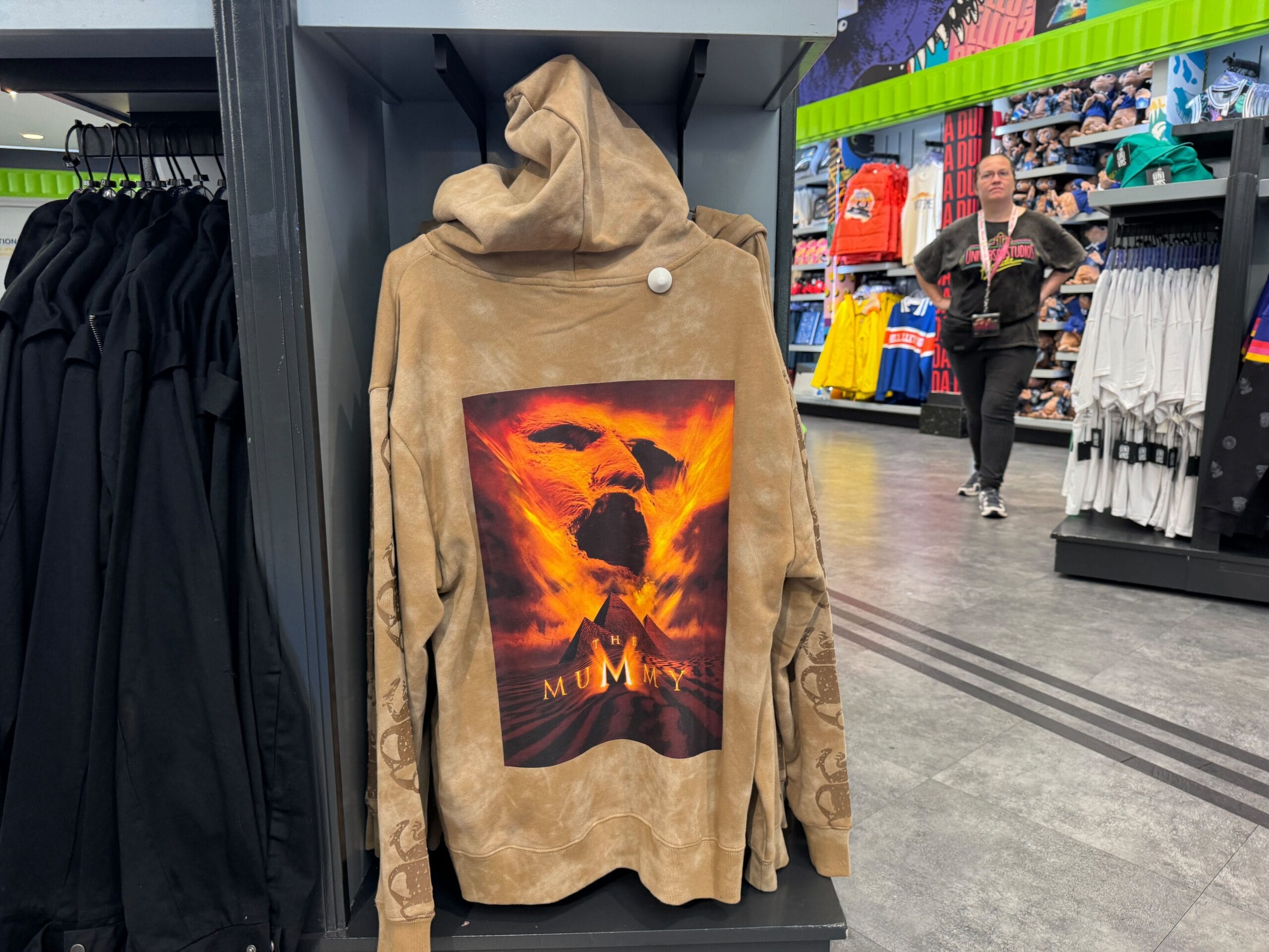 Brown hoodie from the Mummy Collection by UNIVRS on a store rack, with shoppers and clothes in the background.