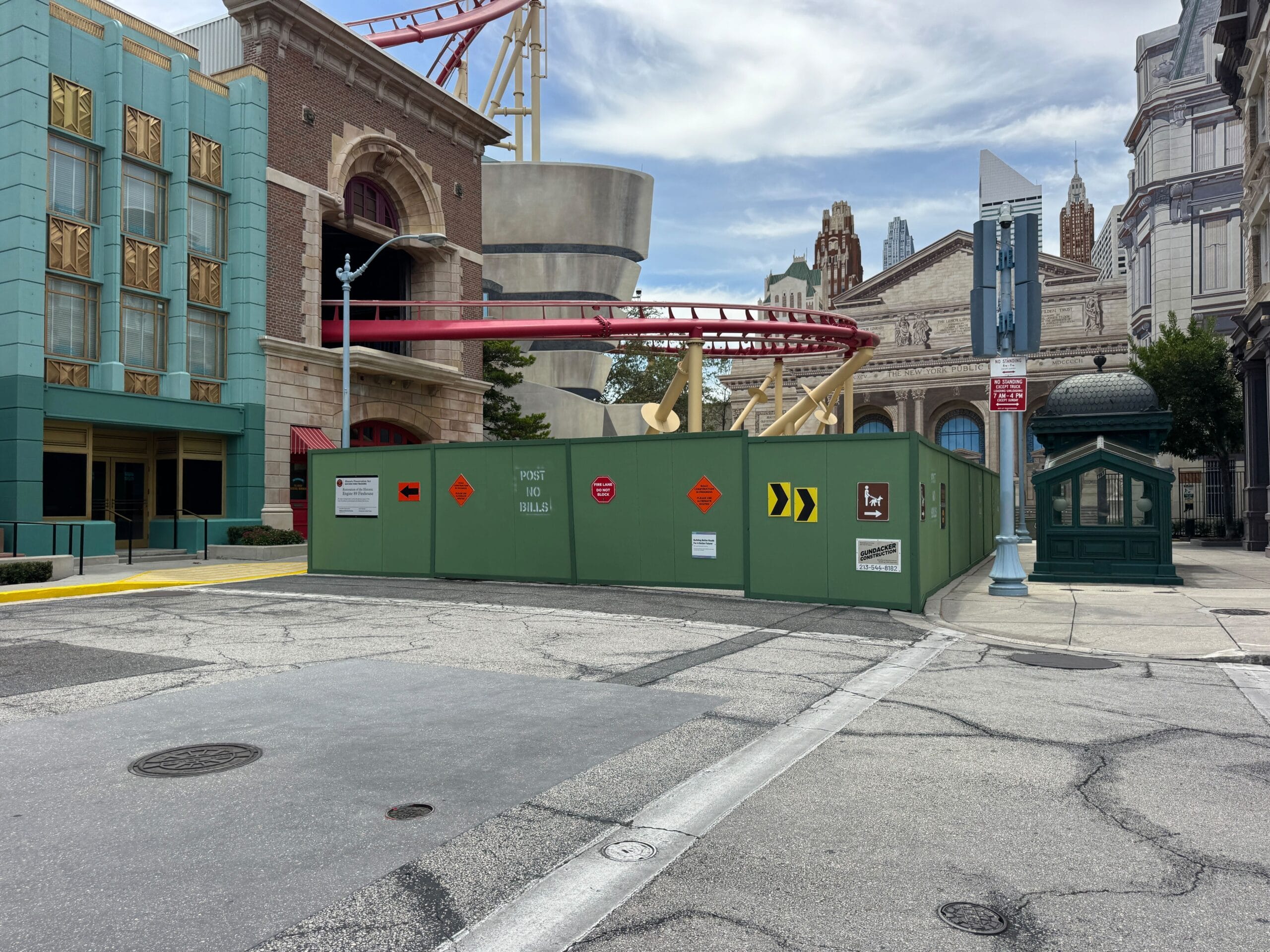 Green construction walls block a city street, with roller coaster tracks and buildings visible in the background.