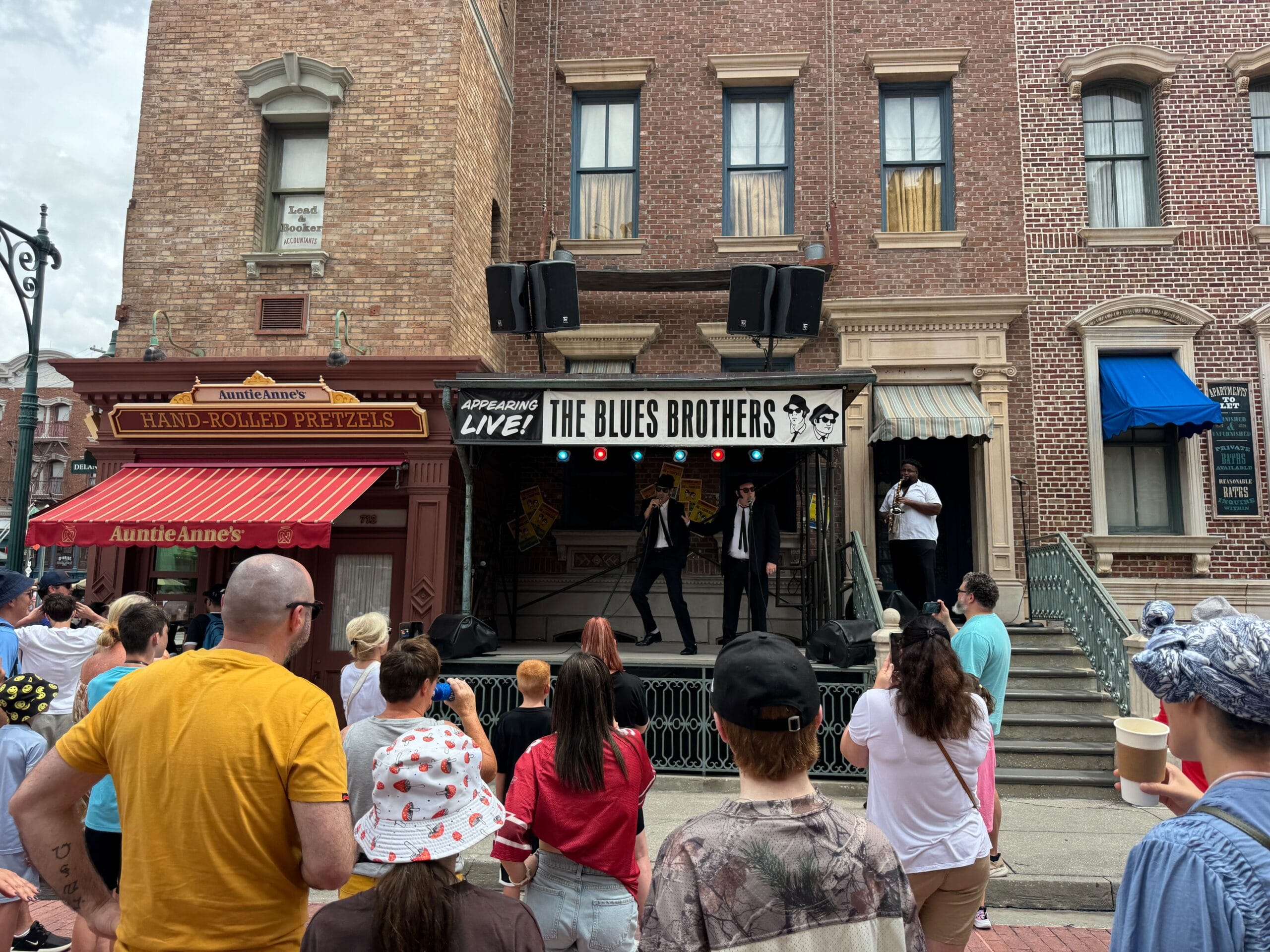 A crowd watches the Blues Brothers perform live near Auntie Anne’s on a lively city street.