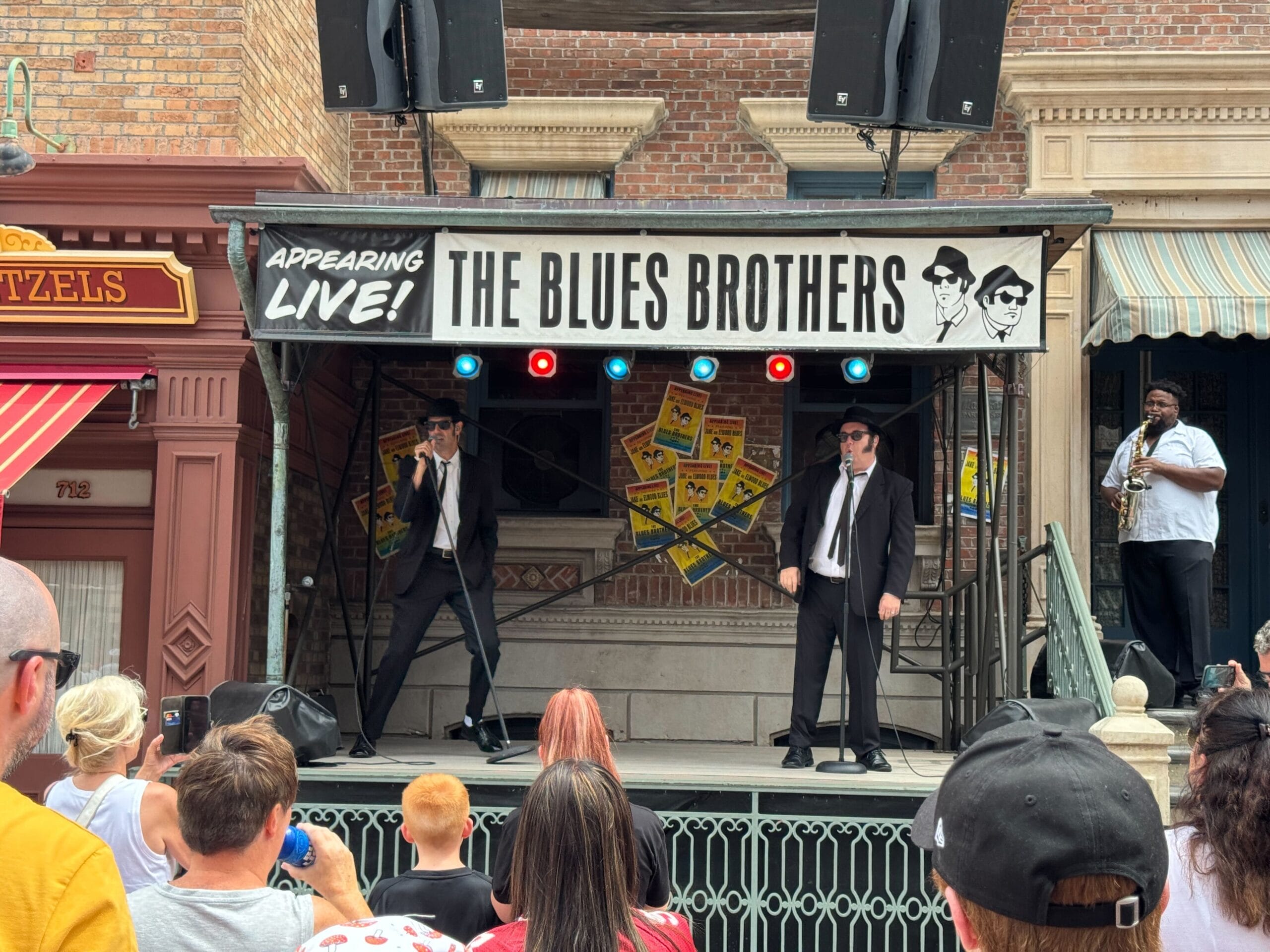 The Blues Brothers perform on stage with a saxophonist; crowd watches outdoors.