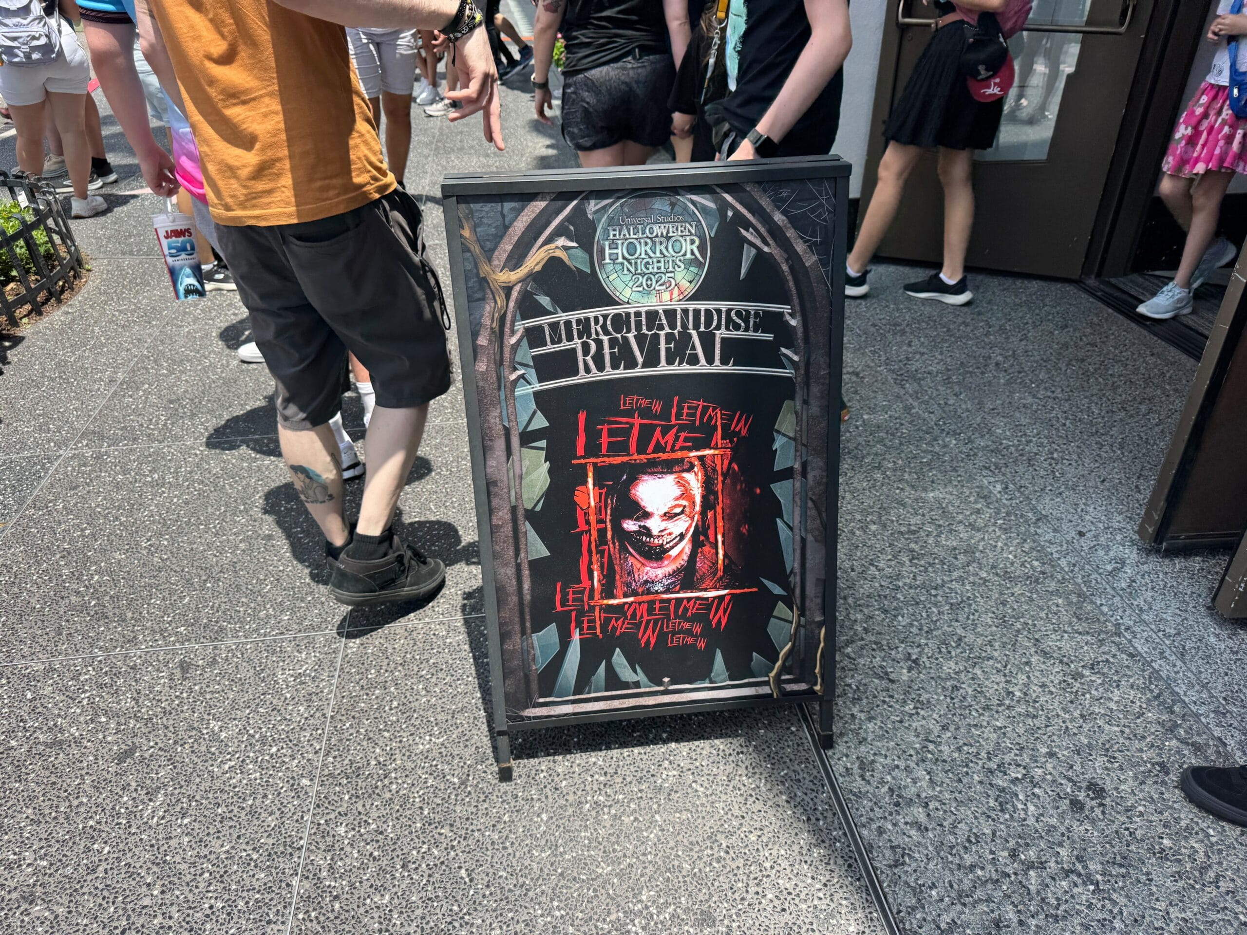 A Halloween Horror Nights merchandise reveal sign with a creepy red face and the words "Let Me In" repeated.
