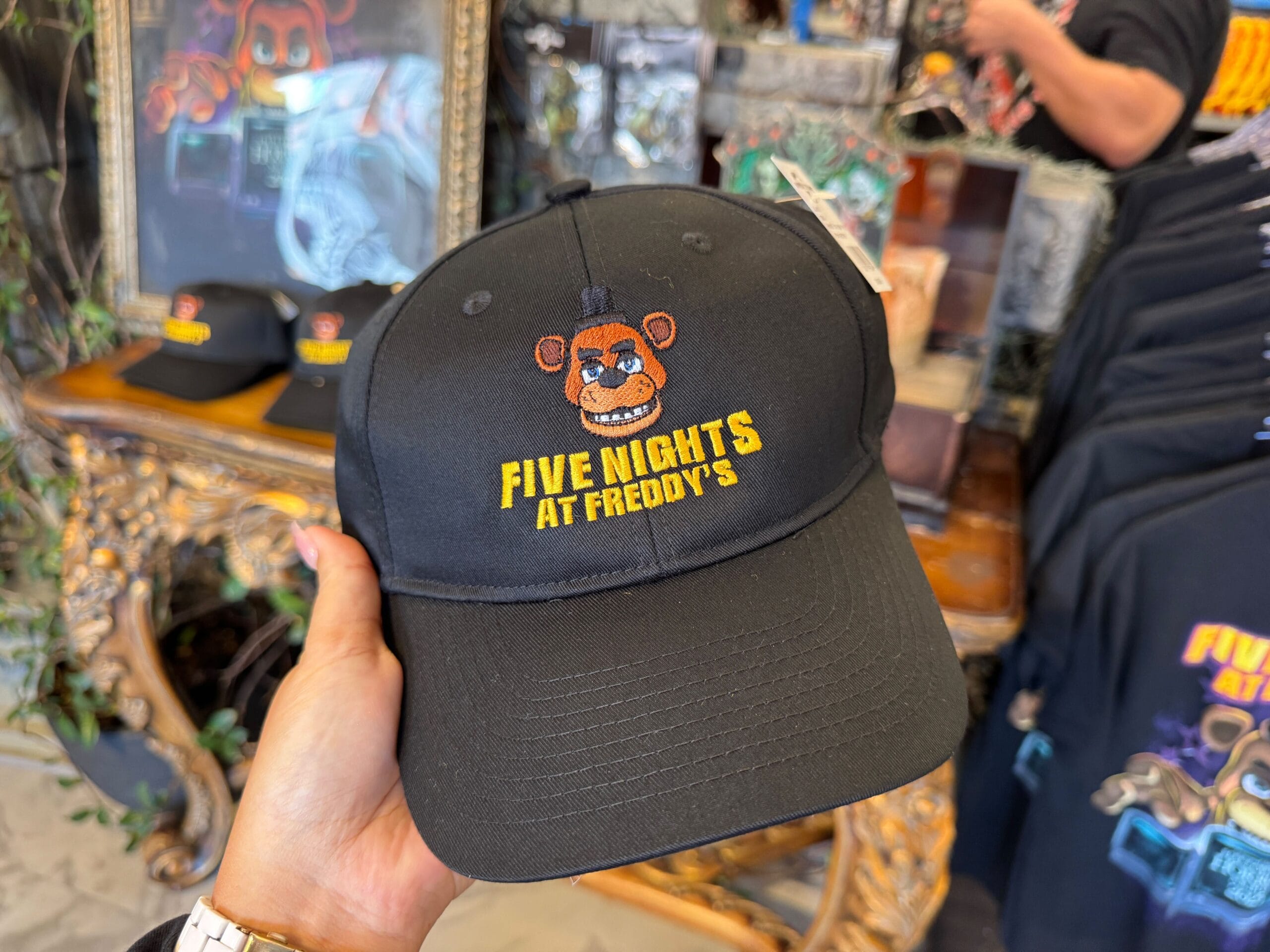 A hand holds a black "Five Nights at Freddy's" cap with Freddy's face embroidered on the front in a store.