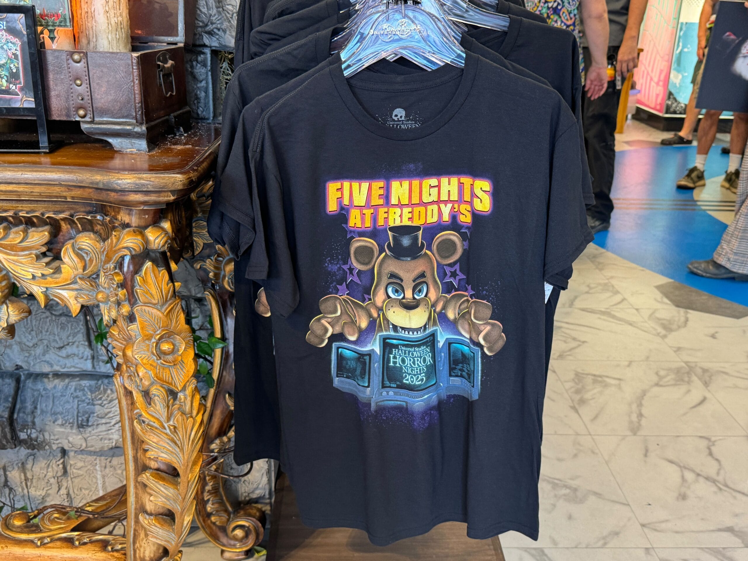 A black "Five Nights at Freddy’s" T-shirt hangs on display in a store with ornate gold décor nearby.