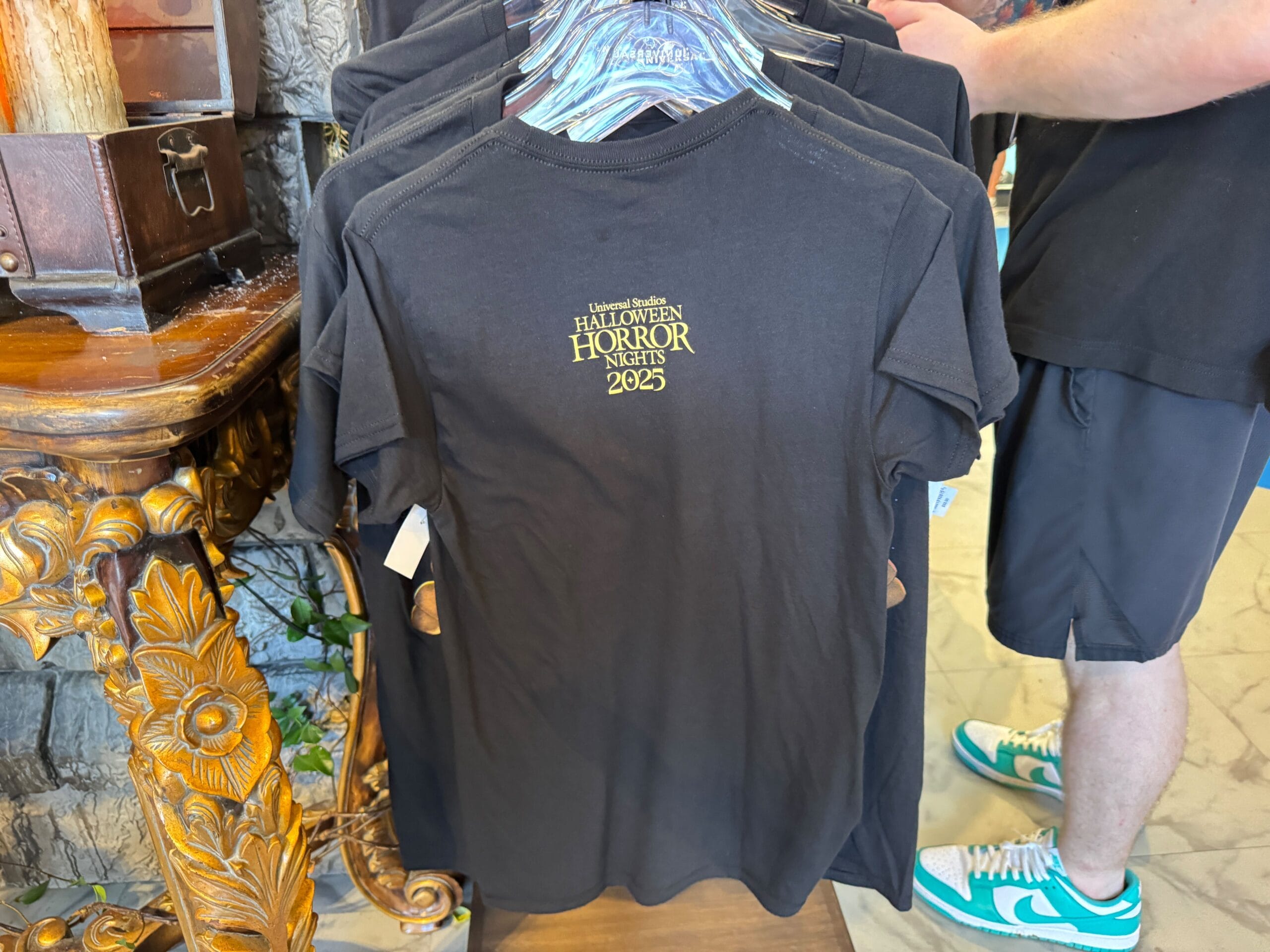 A black t-shirt on a rack with "Halloween Horror Nights 2025" printed in gold on the back.