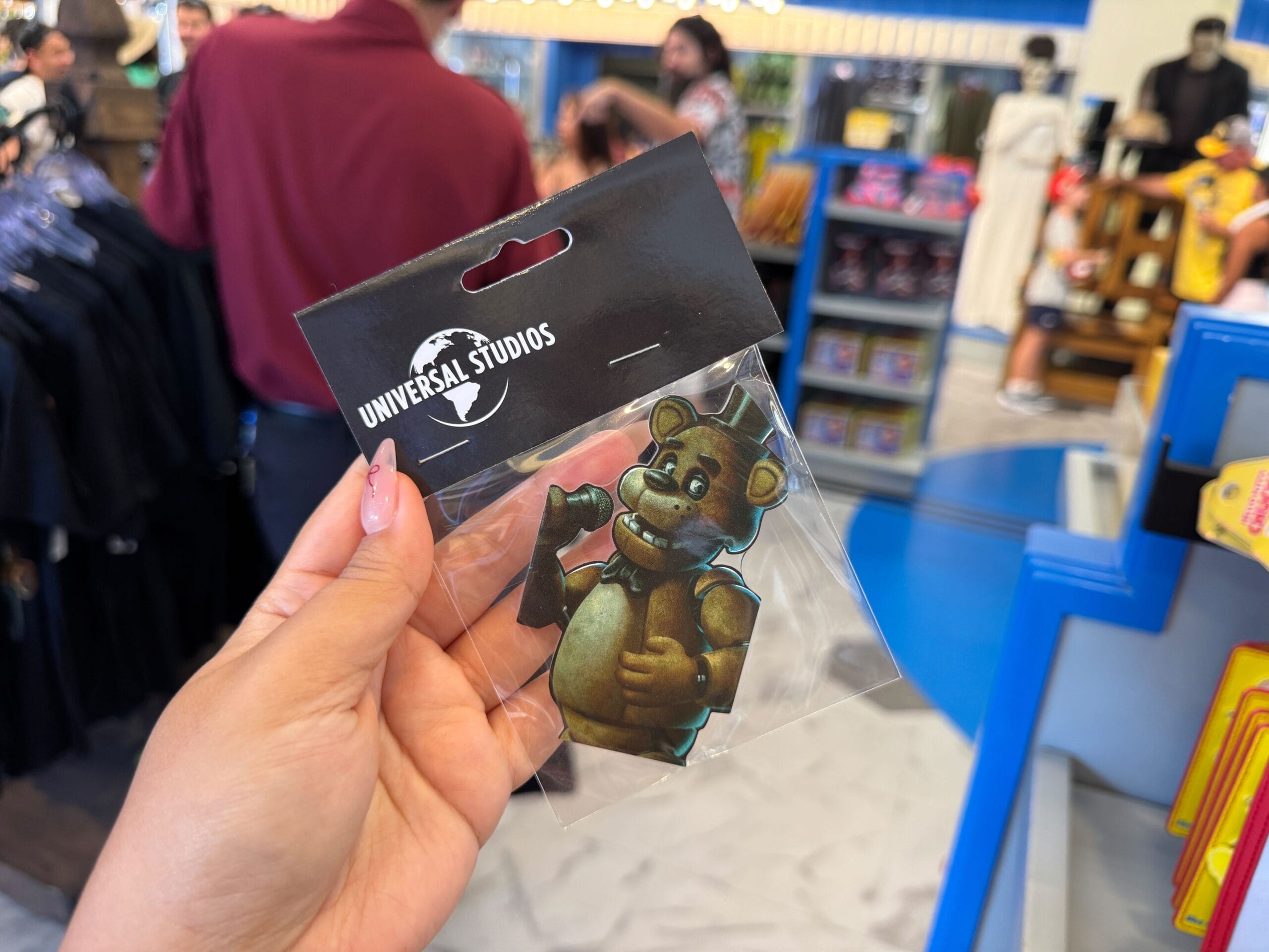A hand holds a Freddy Fazbear Universal Studios magnet in a gift shop with people and merchandise in the background.