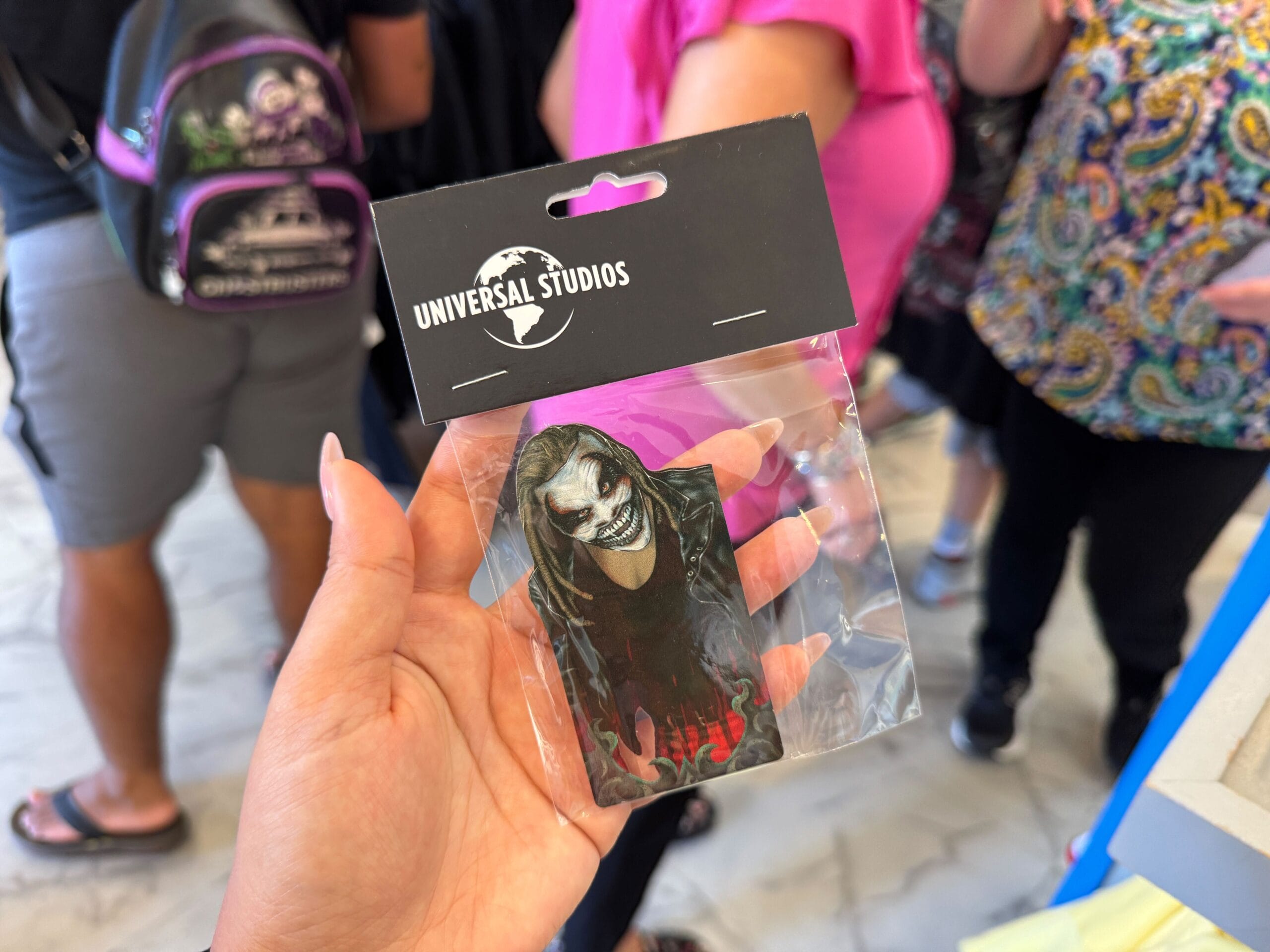 Hand holding a Universal Studios air freshener featuring a character with white face paint and dark makeup in a crowded store.