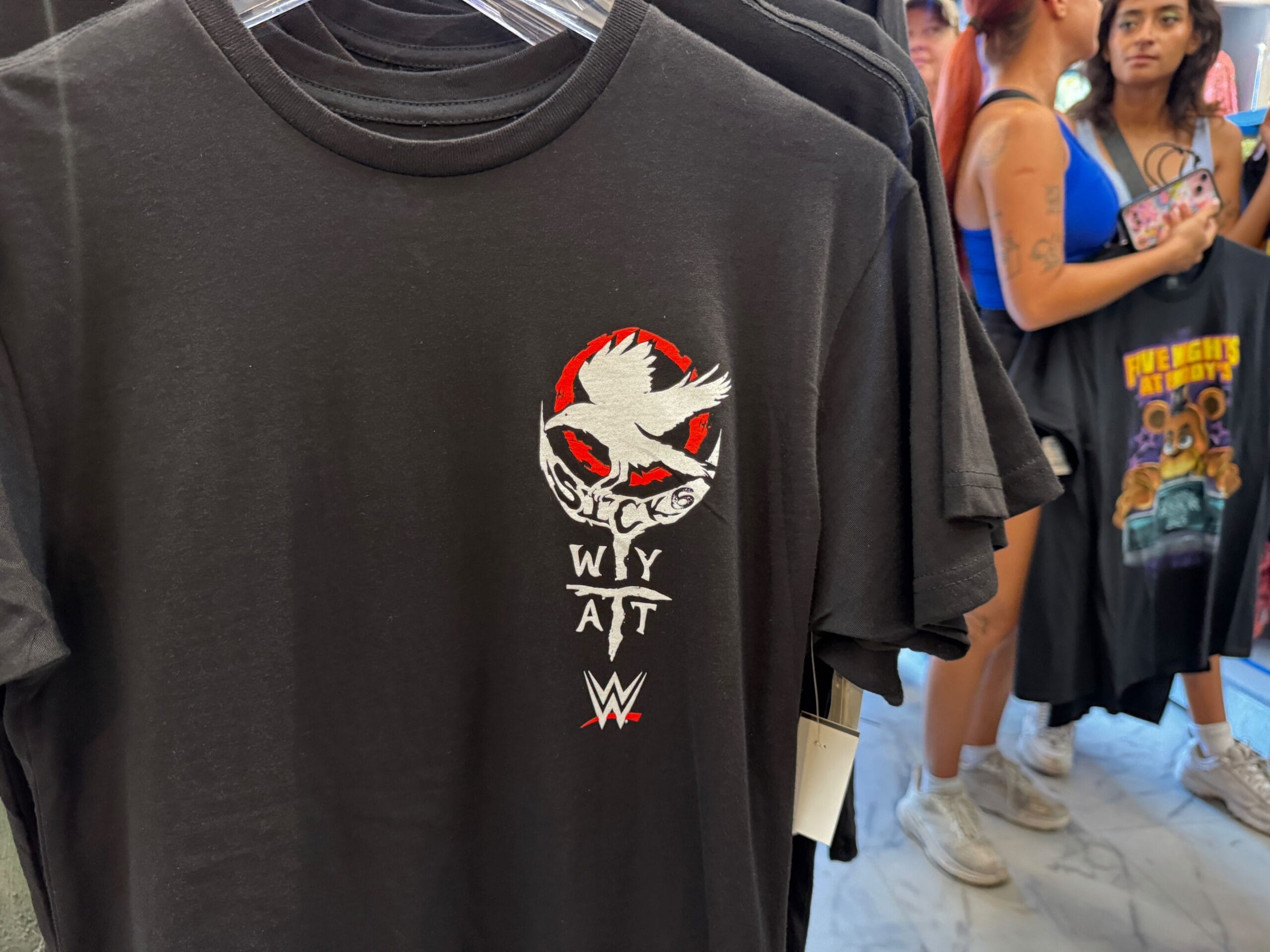 Black WWE t-shirt with a white bird and "Sicks WYA AT" design; people browsing shirts in the background.