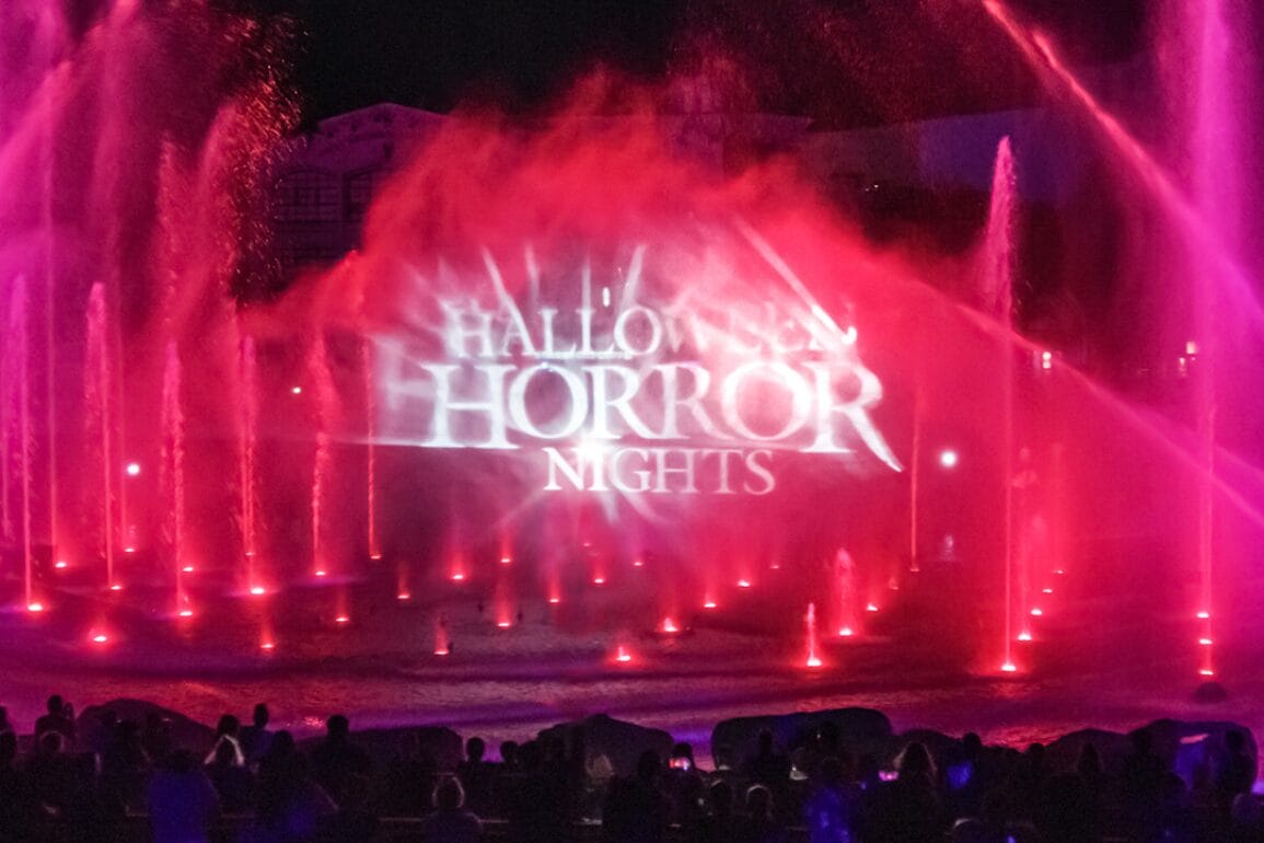 Red and pink fountains with a projected "Halloween Horror Nights" logo, watched by silhouetted crowd at night.
