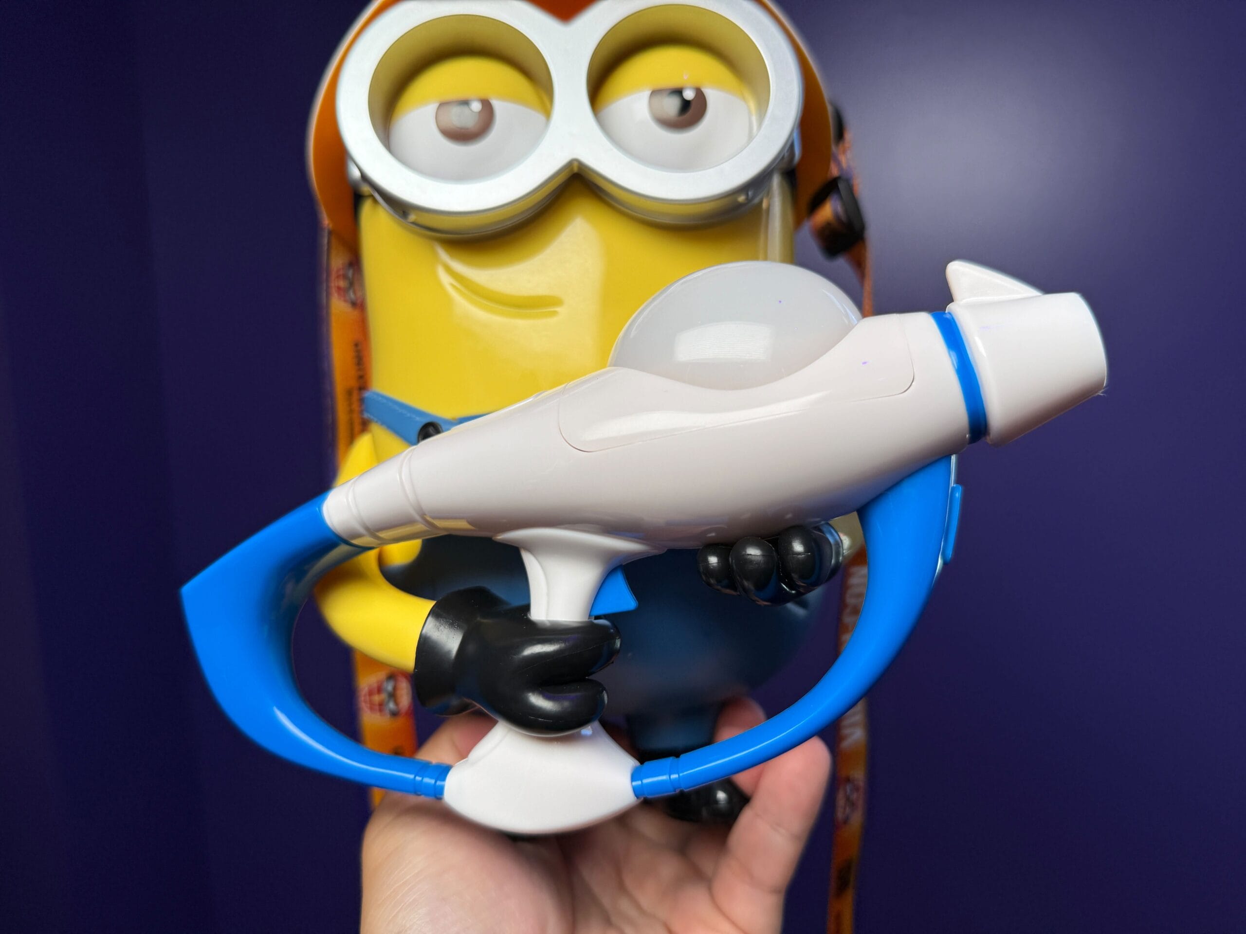 A person holds a Villain-Con Minion Popcorn Bucket with goggles and blaster in front of a purple wall at Universal Studios Japan.