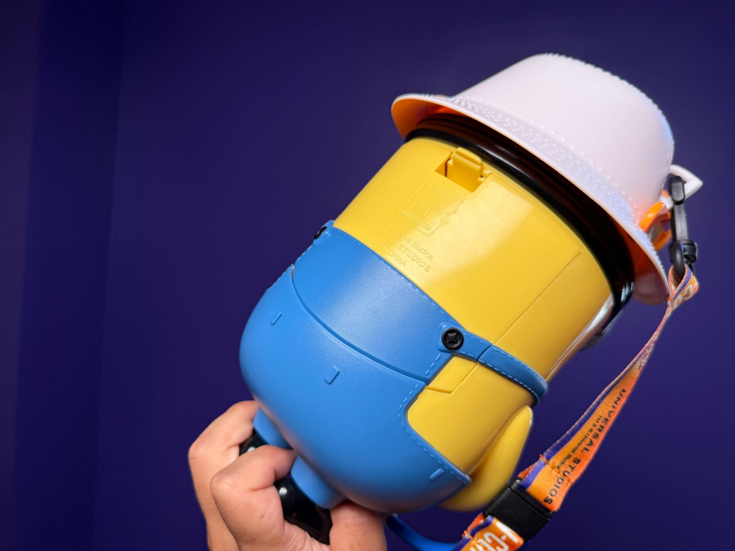 A hand holds a Minion Popcorn Bucket wearing a white construction helmet against a purple background at Universal Studios Japan.