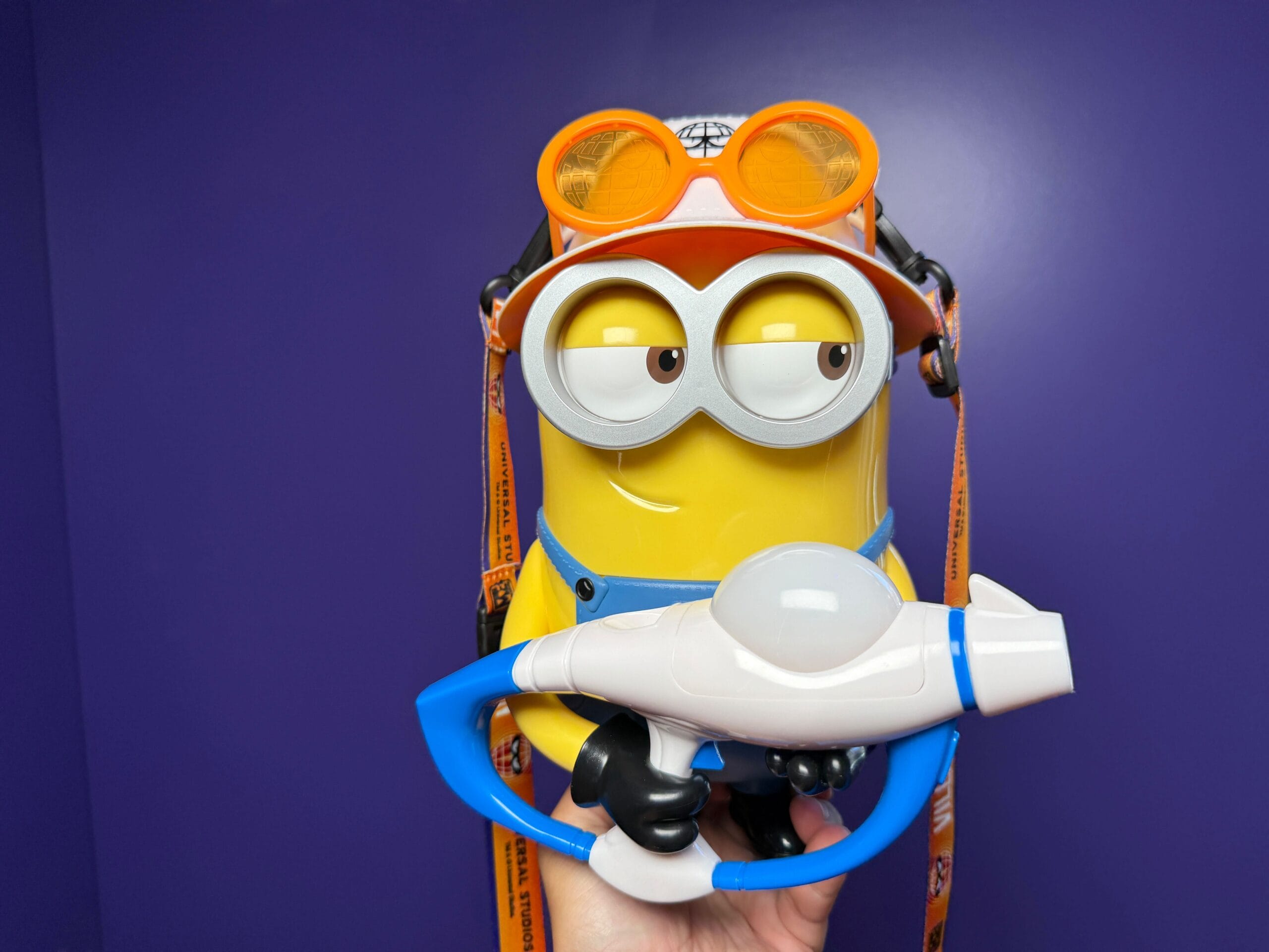 A hand holds a Minion Popcorn Bucket wearing orange goggles and holding a blue blaster against a purple background.