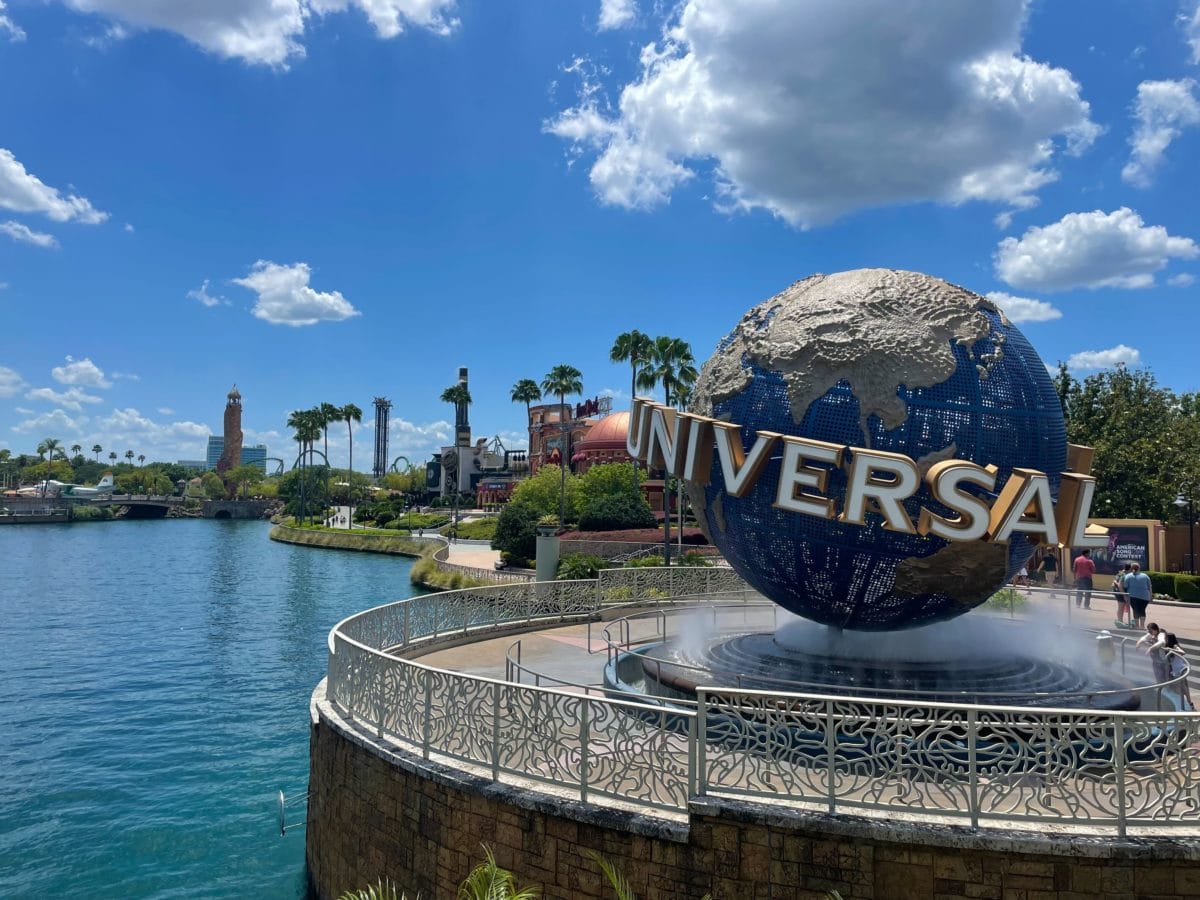 Large globe with "Universal" logo at Universal Orlando Resort entrance, blue sky, water, and team members in the background.