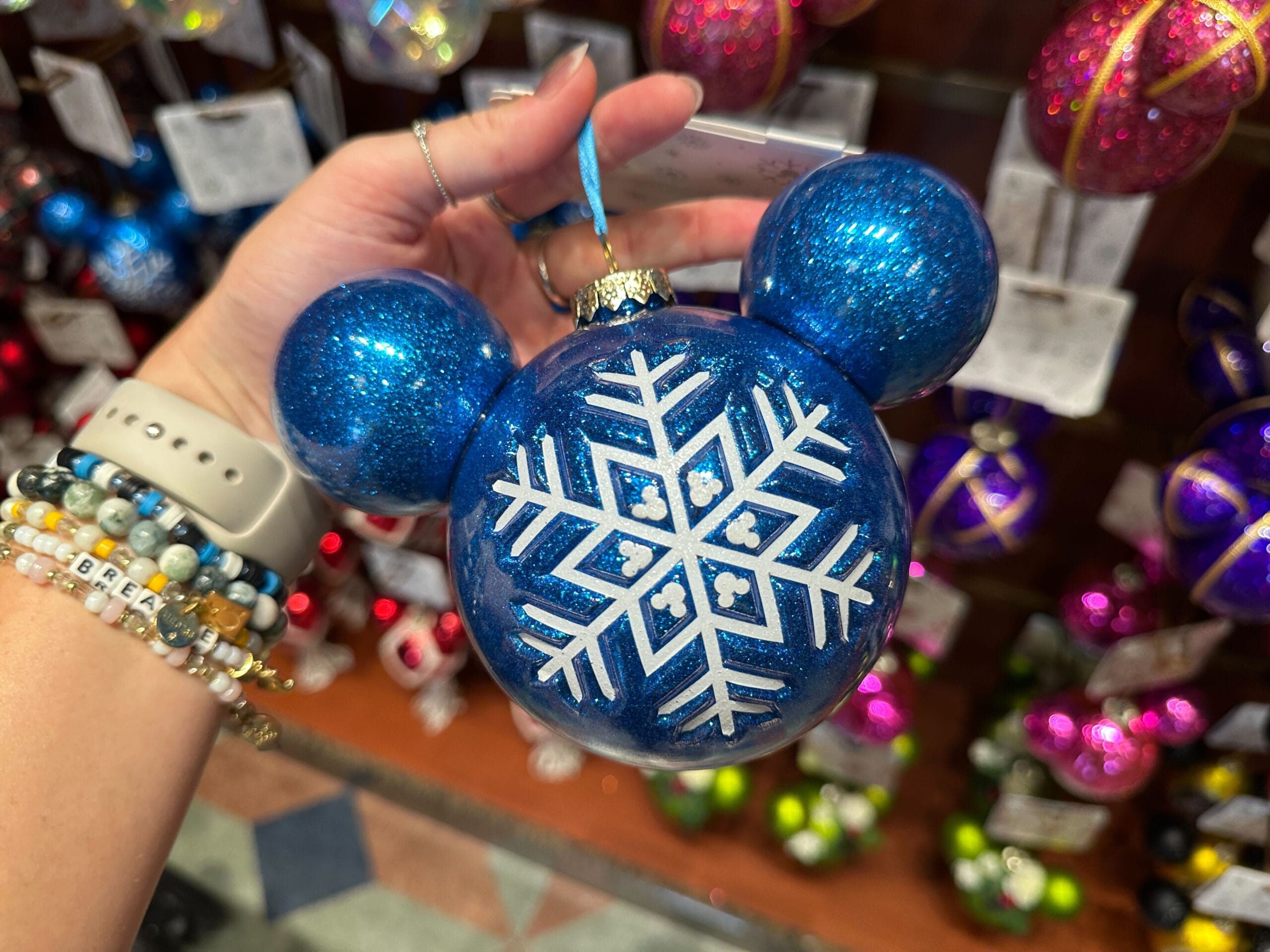 A hand holds a blue Mickey Mouse-shaped ornament with a white snowflake design in front of a display of ornaments.