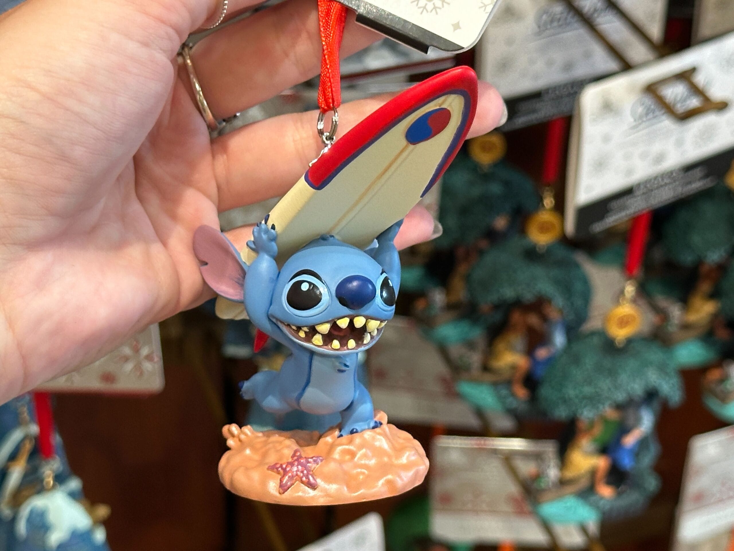 A hand holds a Stitch ornament with a surfboard, smiling and standing on sand with a starfish.
