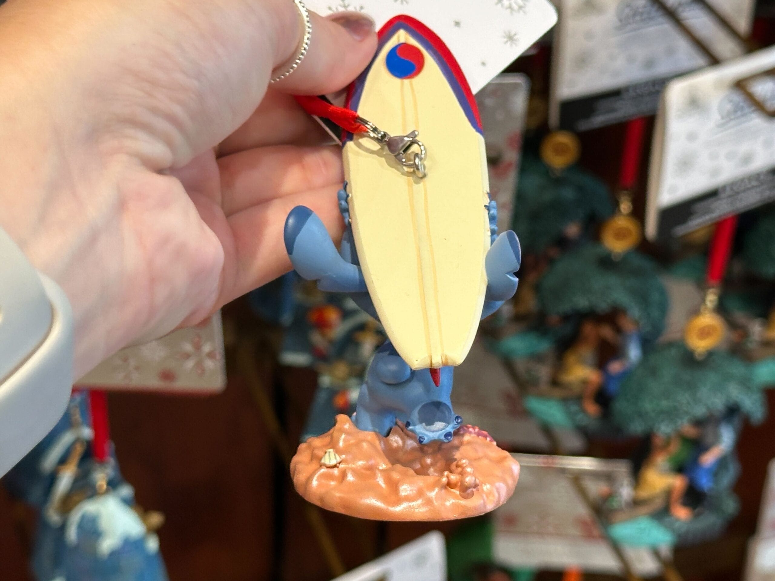 A hand holds a blue creature figurine with a surfboard, displayed among other hanging ornaments.