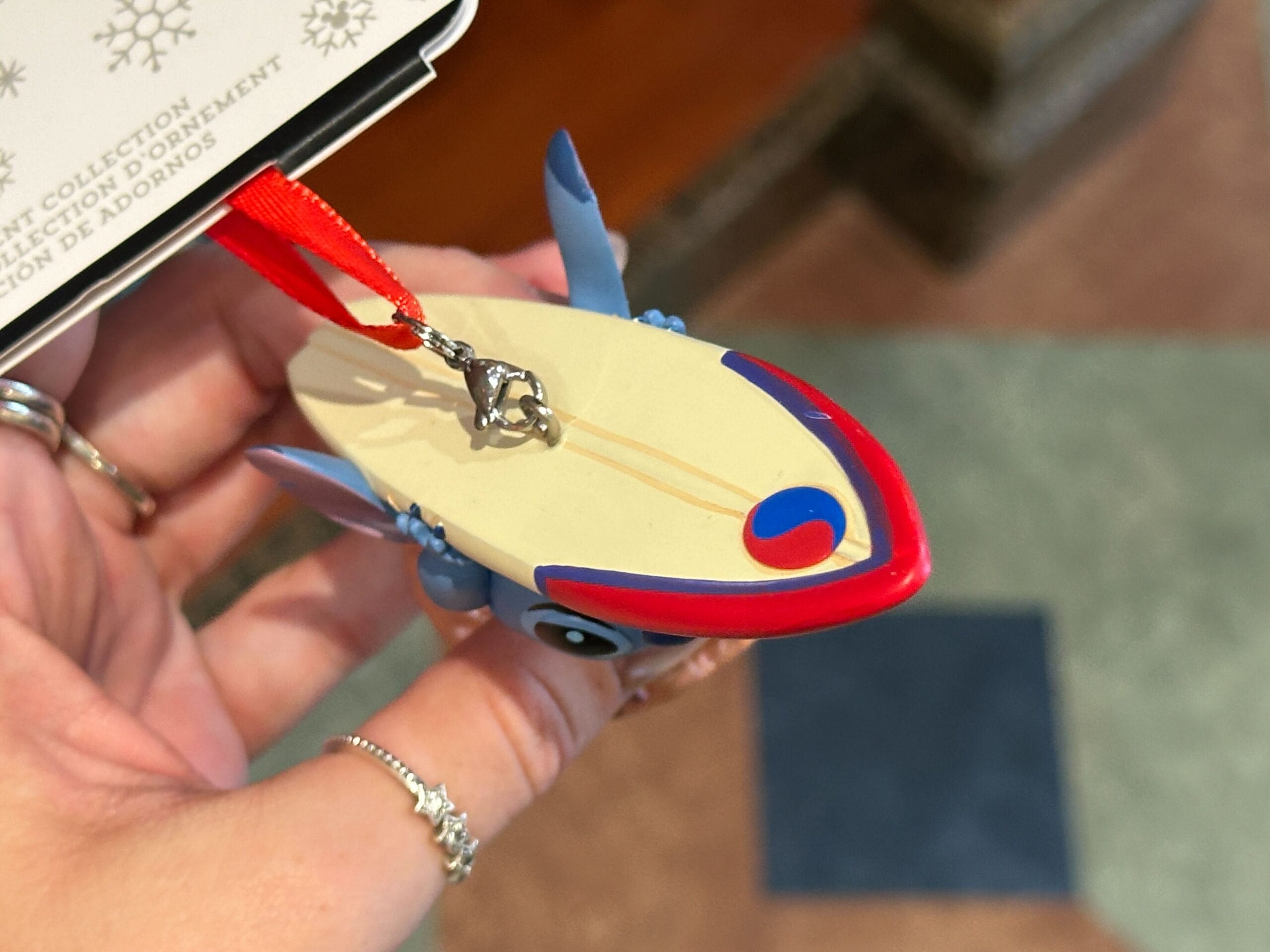 A hand holds a Stitch ornament riding a surfboard with a red ribbon and silver hook.