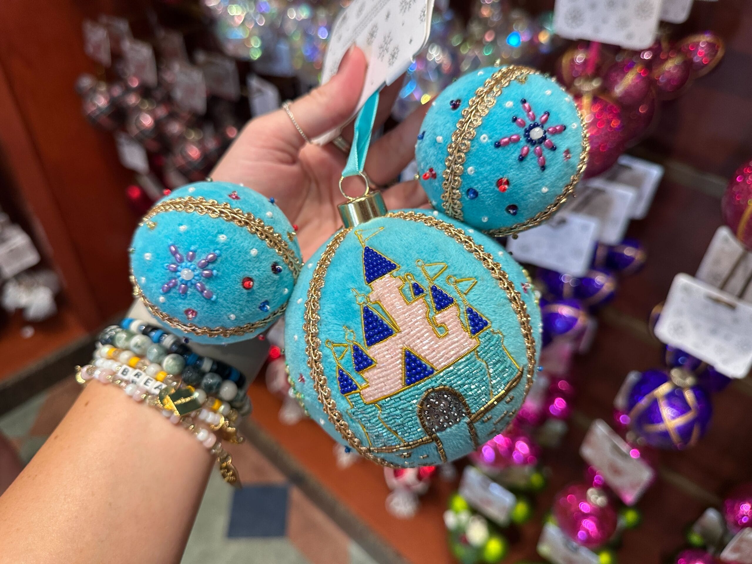 A hand holds a blue, Mickey-shaped ornament with a castle design, surrounded by other colorful ornaments.