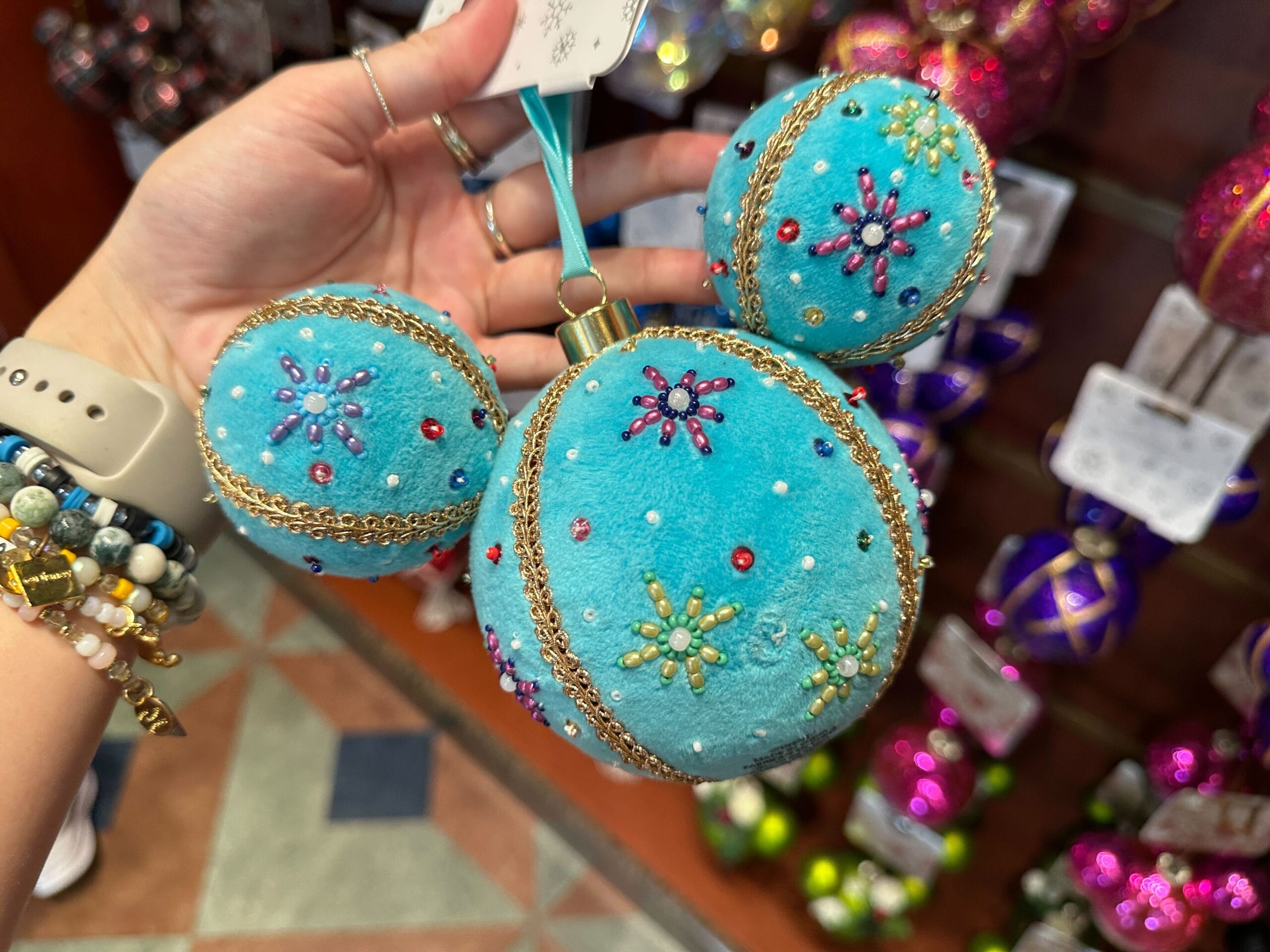 Hand holding a blue Mickey Mouse-shaped ornament decorated with beads and gold trim, surrounded by other ornaments.