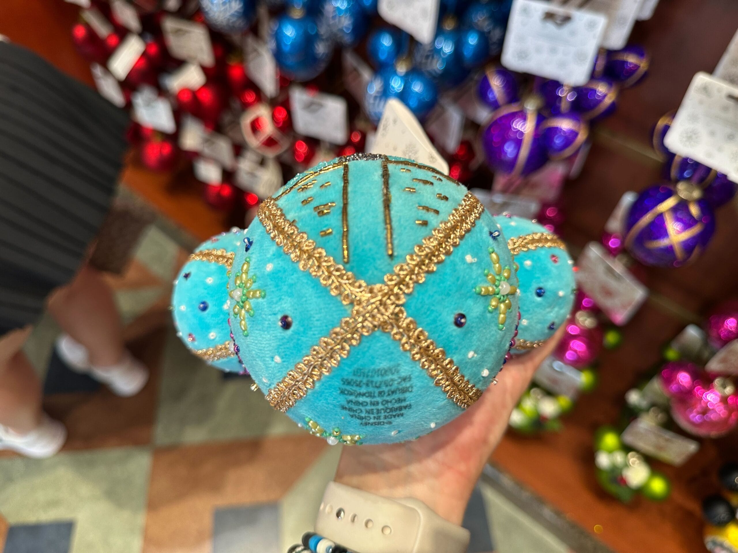 A hand holds a blue and gold bejeweled ornament, with colorful ornaments displayed on shelves in the background.