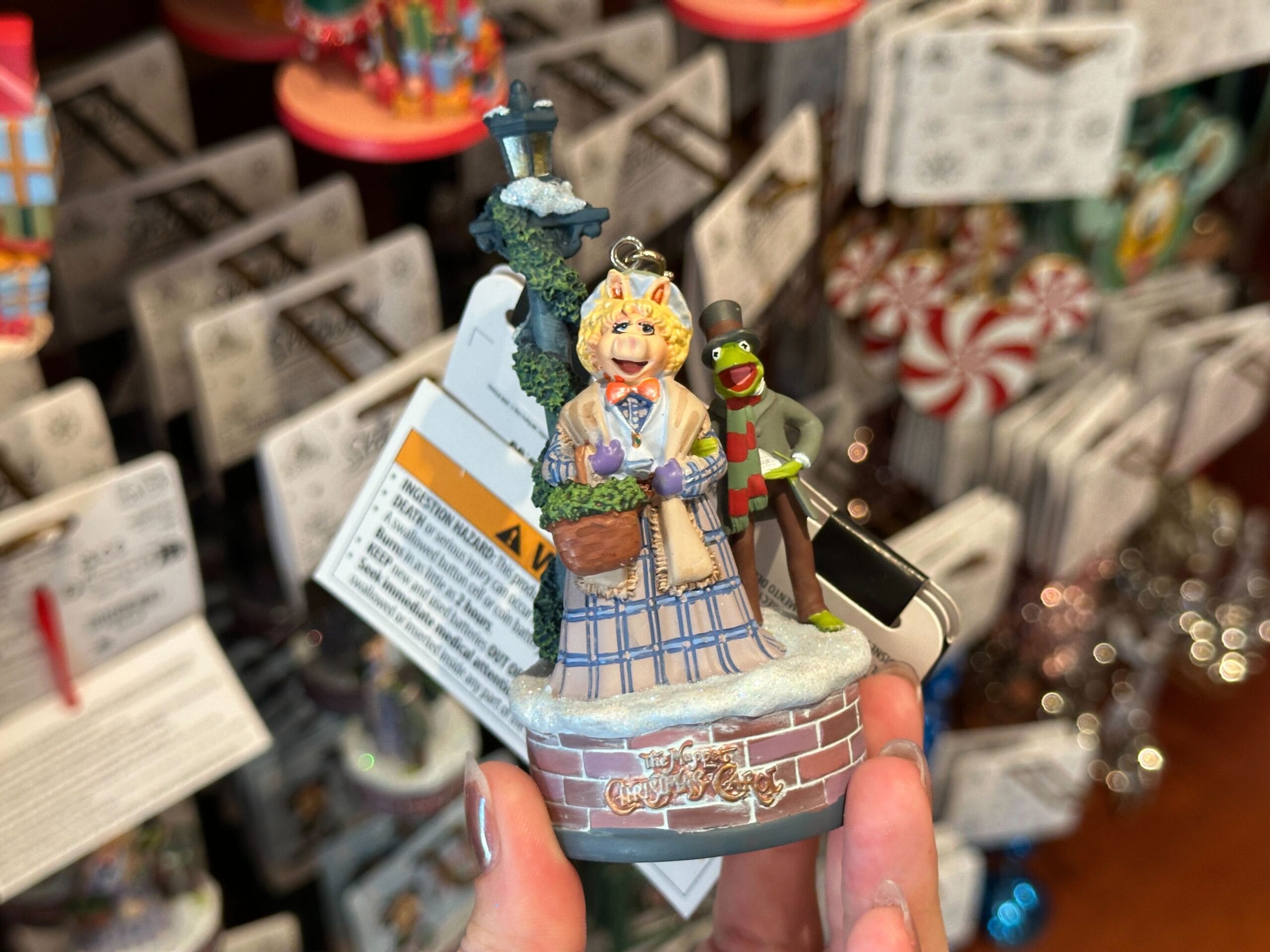 Hand holding a Muppet Christmas Carol ornament featuring Miss Piggy and Kermit, with labels visible in the background.