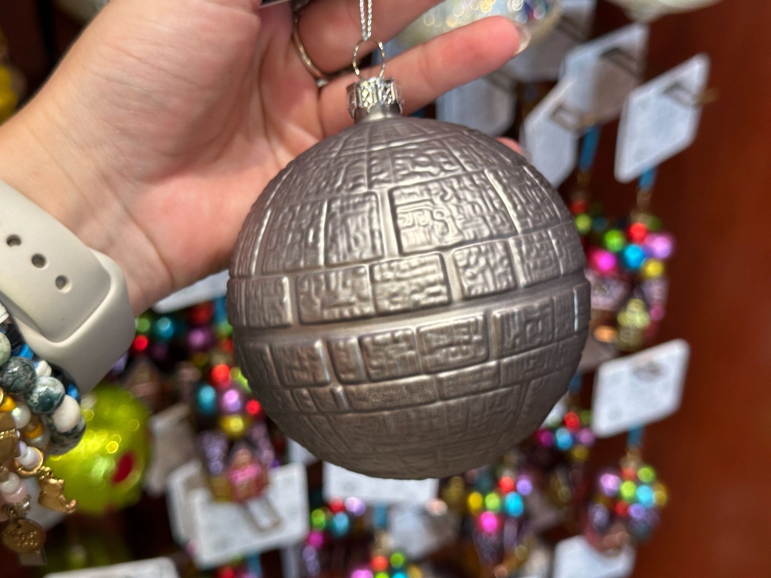 A hand holds a gray, Death Star-shaped ornament with colorful ornaments blurred in the background.