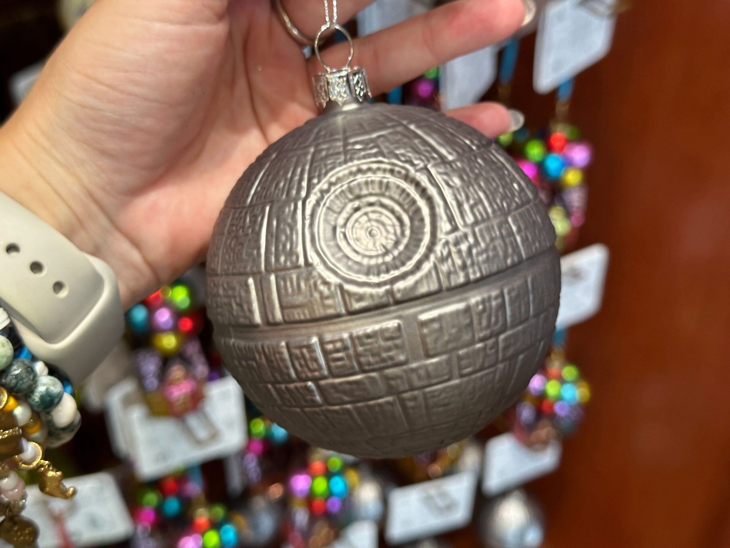 A hand holds a Death Star-shaped Christmas ornament with colorful ornaments blurred in the background.