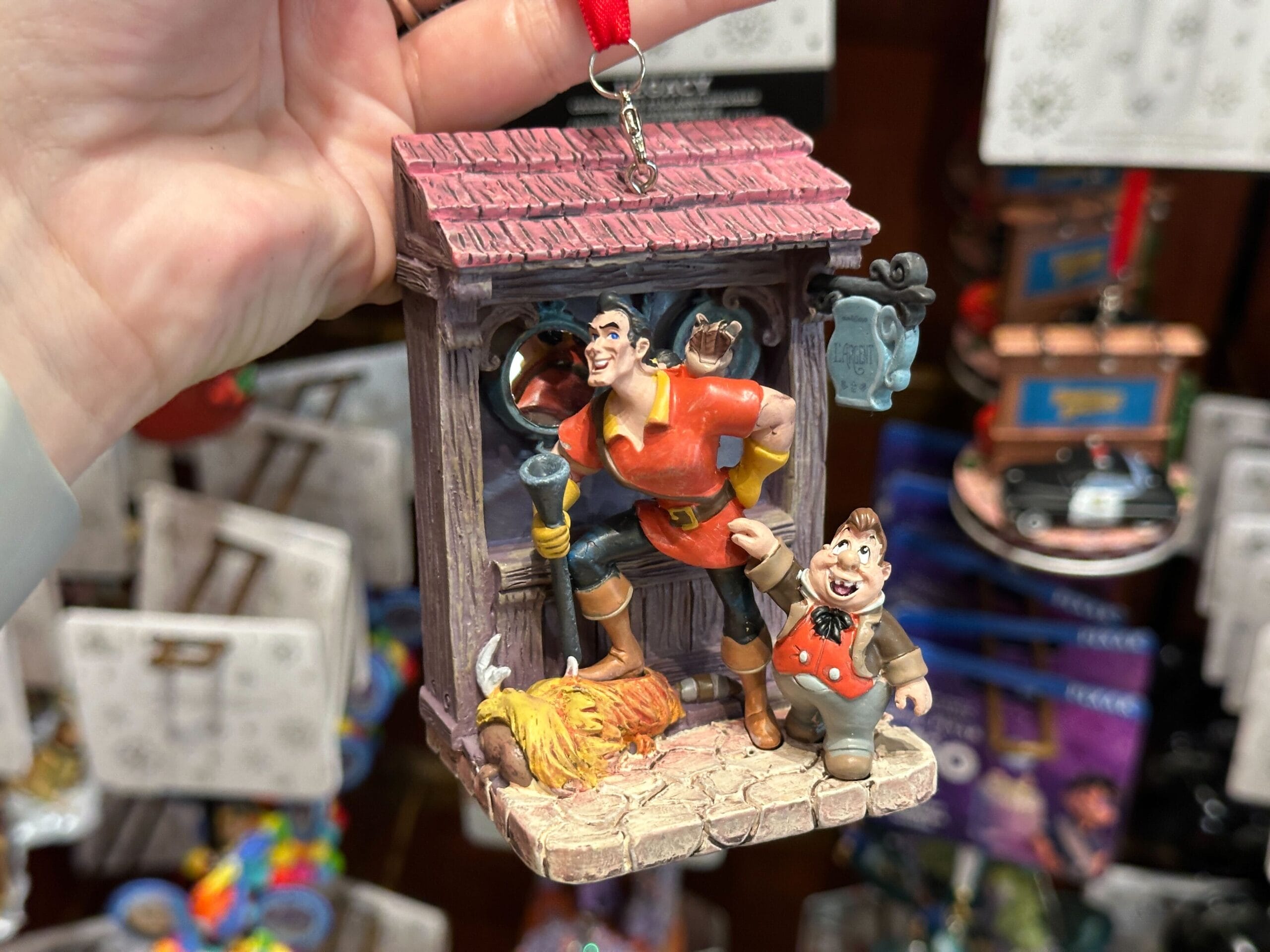 A hand holds a Gaston and LeFou ornament from "Beauty and the Beast" in front of a display of other ornaments.