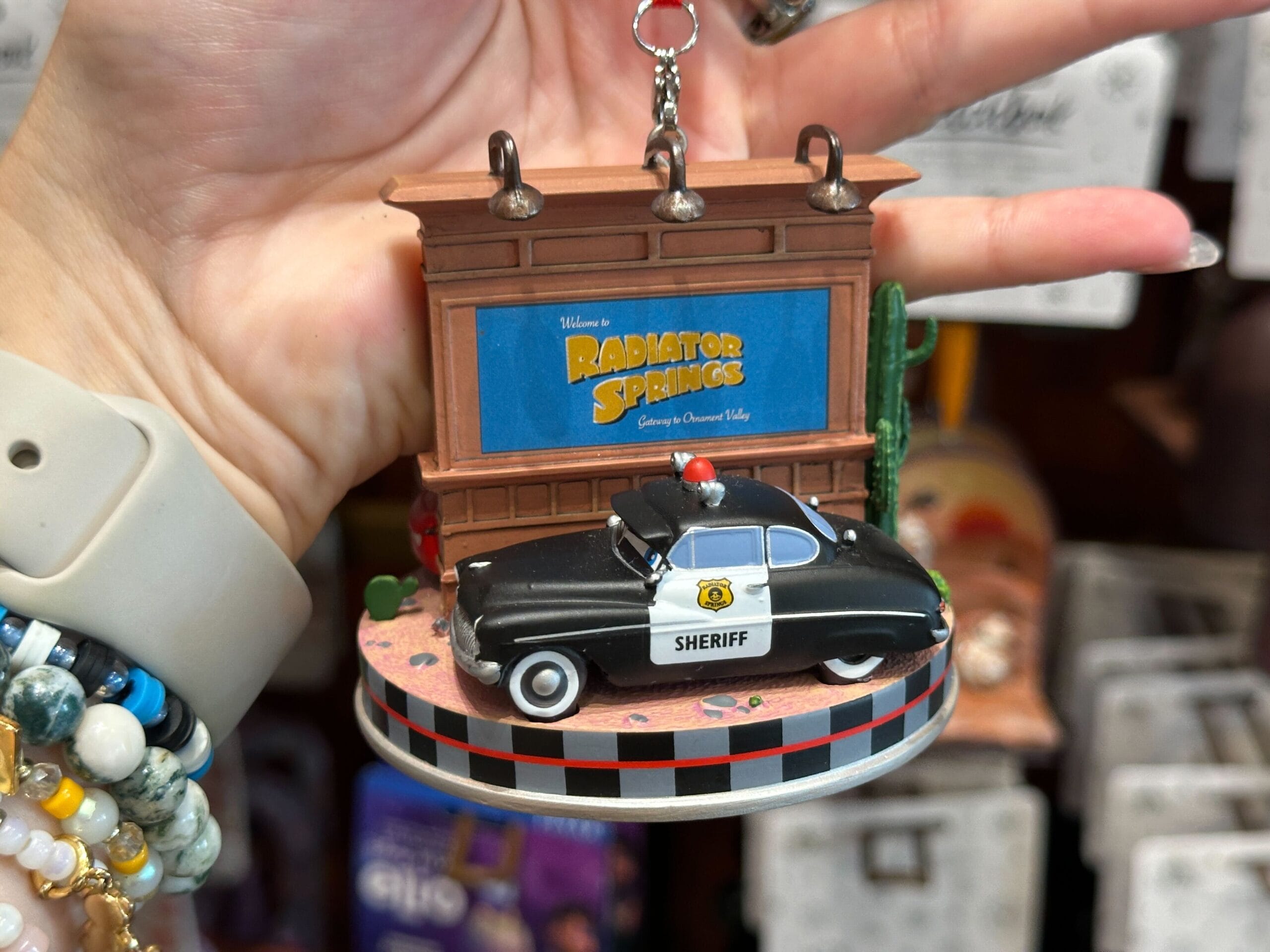 Hand holding a Radiator Springs sheriff car ornament in front of a sign, with bracelets on the wrist.
