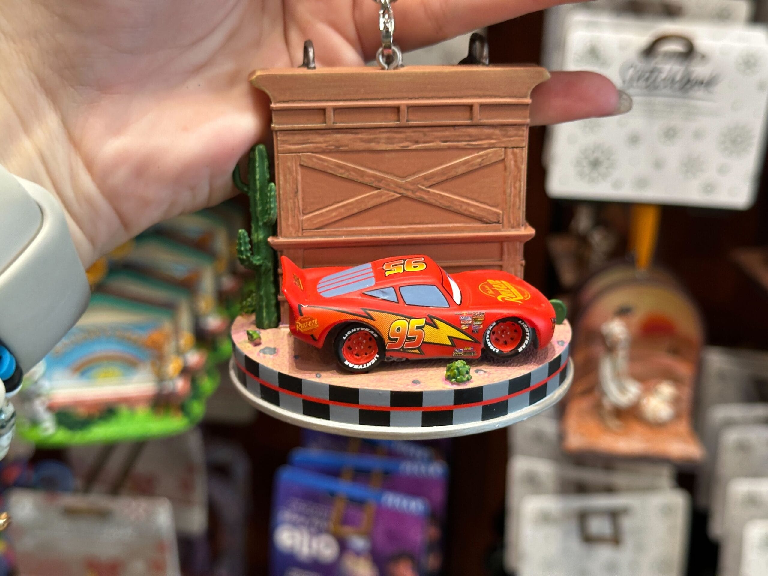 Hand holding a Lightning McQueen ornament from Cars, with a desert scene and building backdrop in a store display.