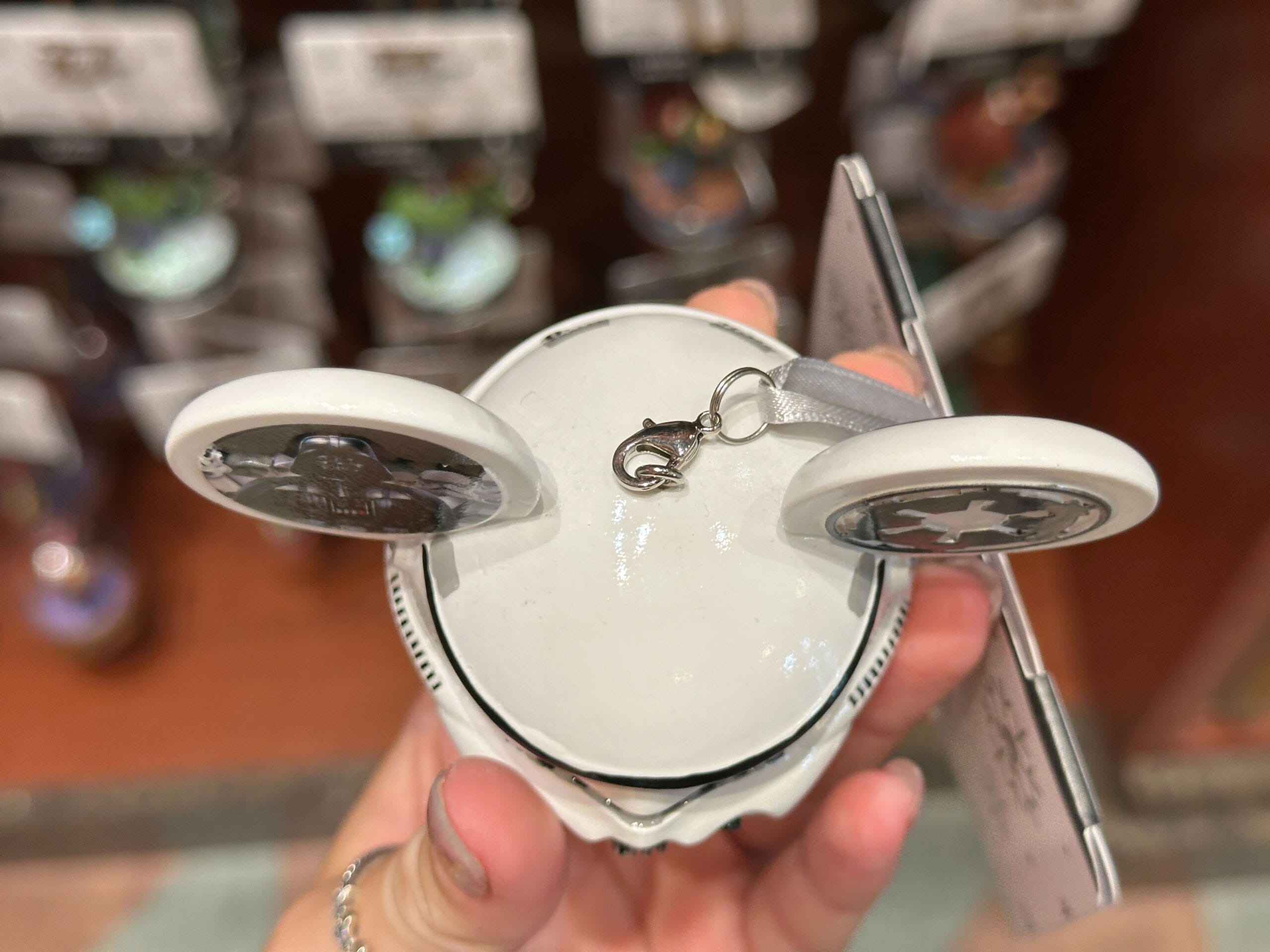 A hand holds a round white ornament with two open lids and a small hanging clasp at the top.