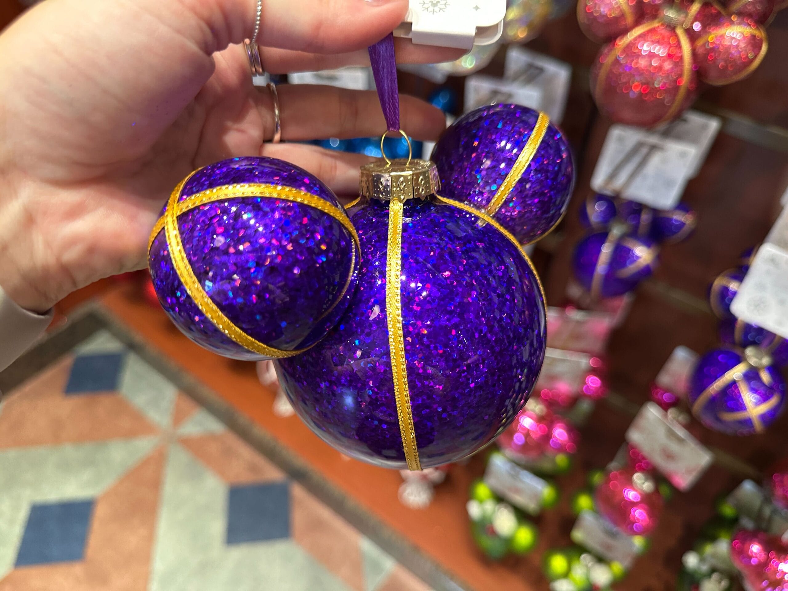 A hand holding a sparkly purple Mickey Mouse-shaped ornament with gold ribbon accents.