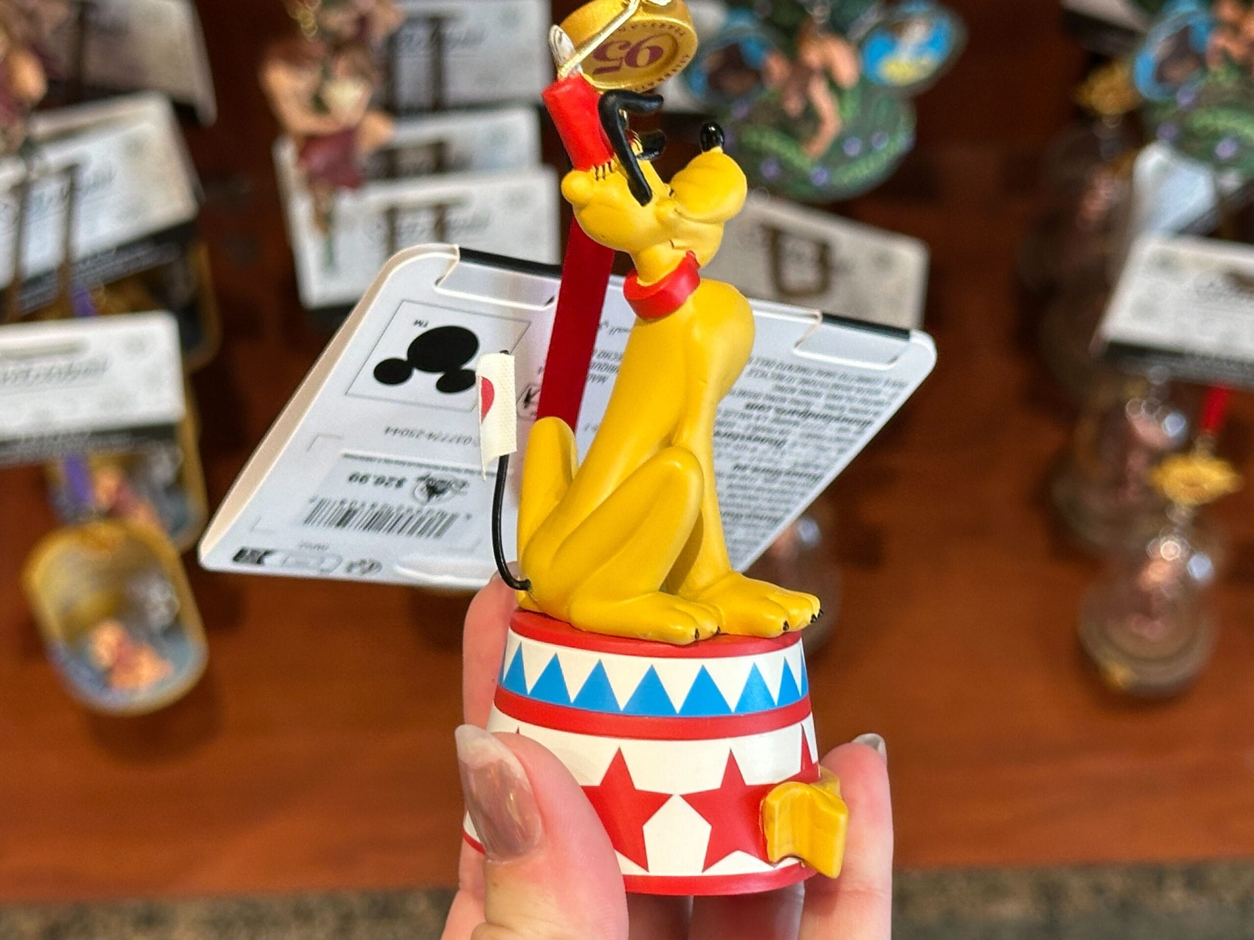 A hand holds a Pluto figurine sitting on a circus platform, with Disney merchandise blurred in the background.