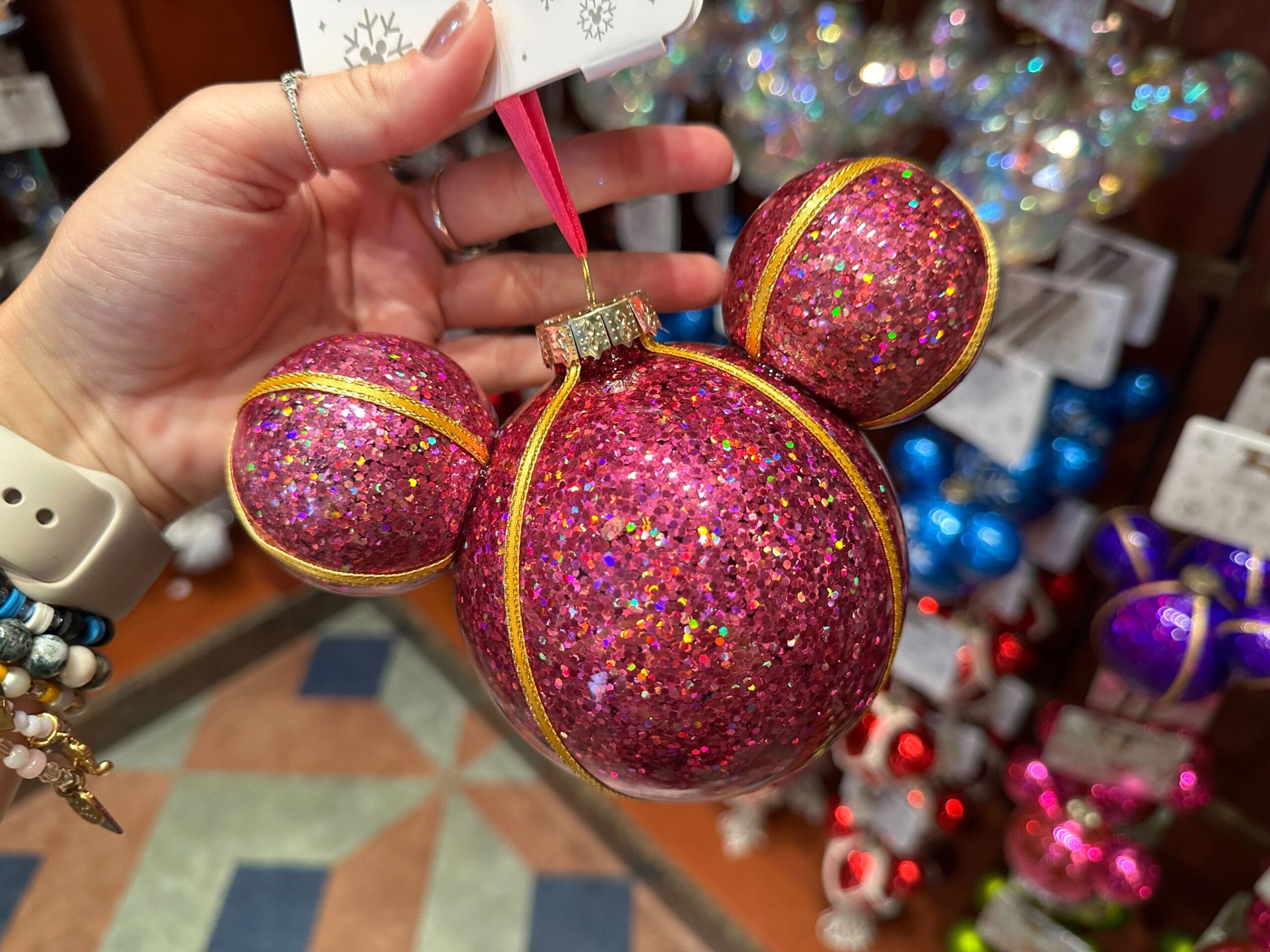 A hand holds a pink, glittery Mickey Mouse-shaped Christmas ornament with gold trim in a store.