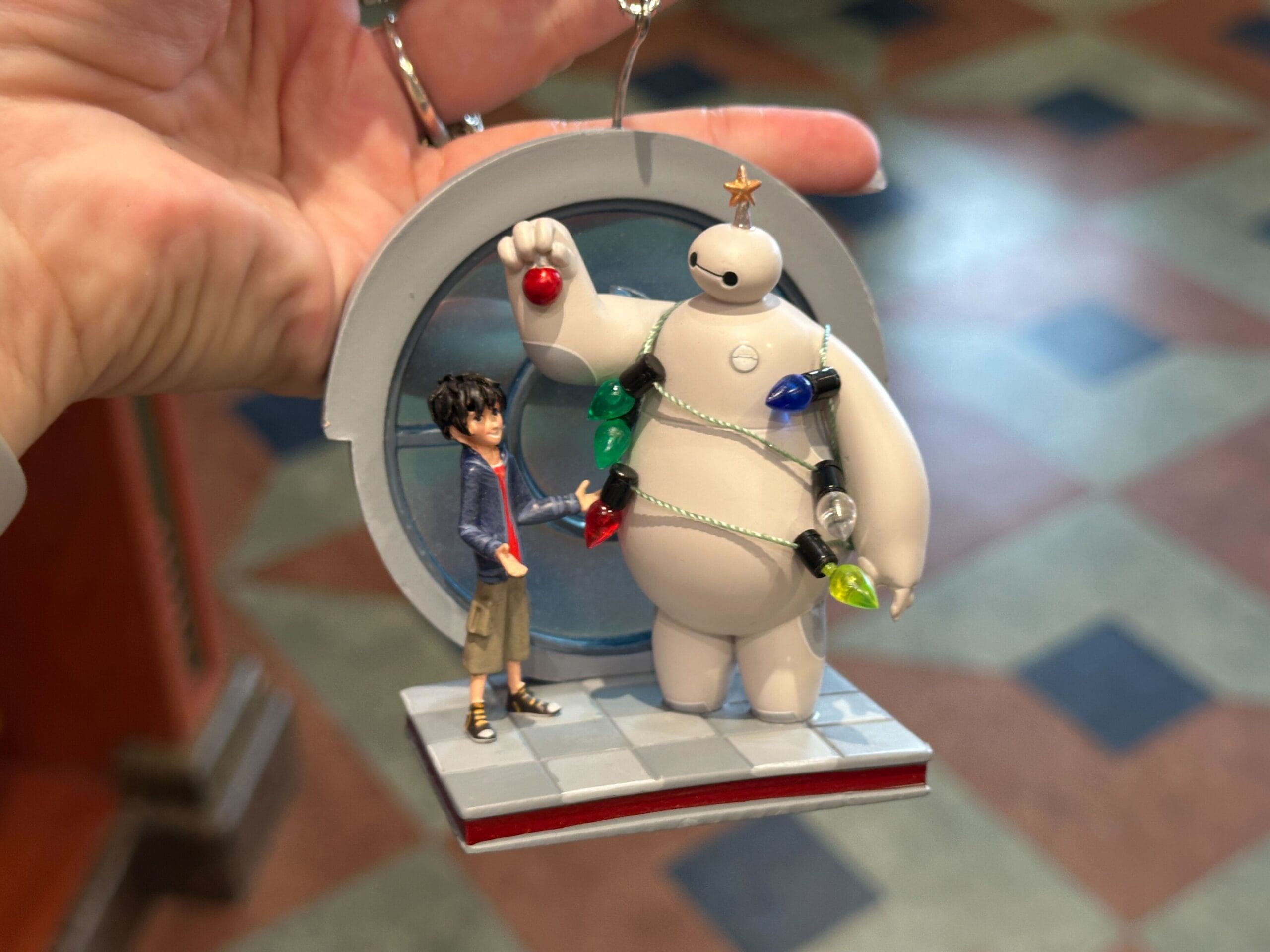 A hand holds an ornament of Hiro and Baymax from Big Hero 6; Baymax is decorated with Christmas lights.