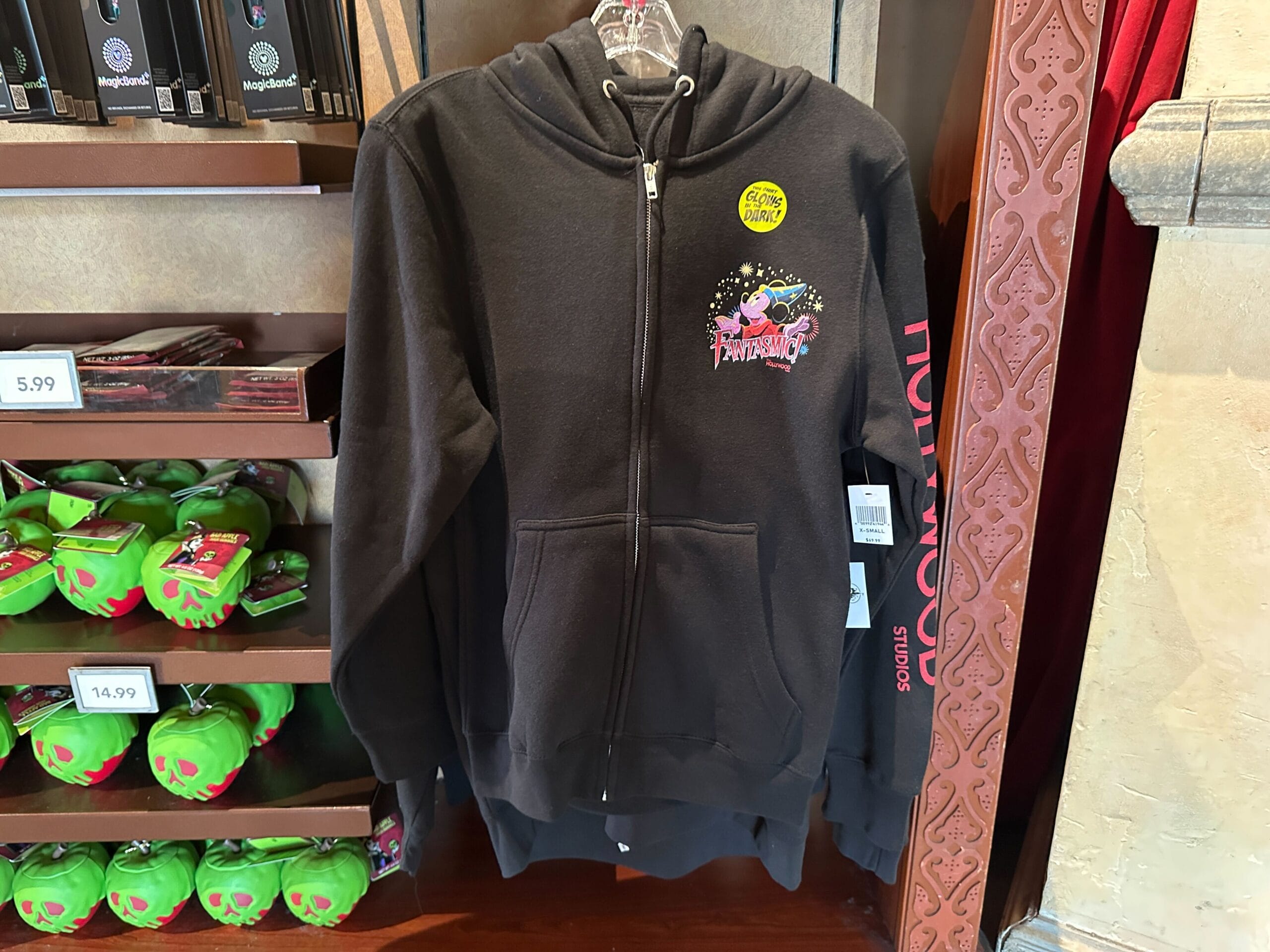 Black Hollywood Studios hoodie with front graphic, zipped, on display in a store next to green apple-themed merchandise.