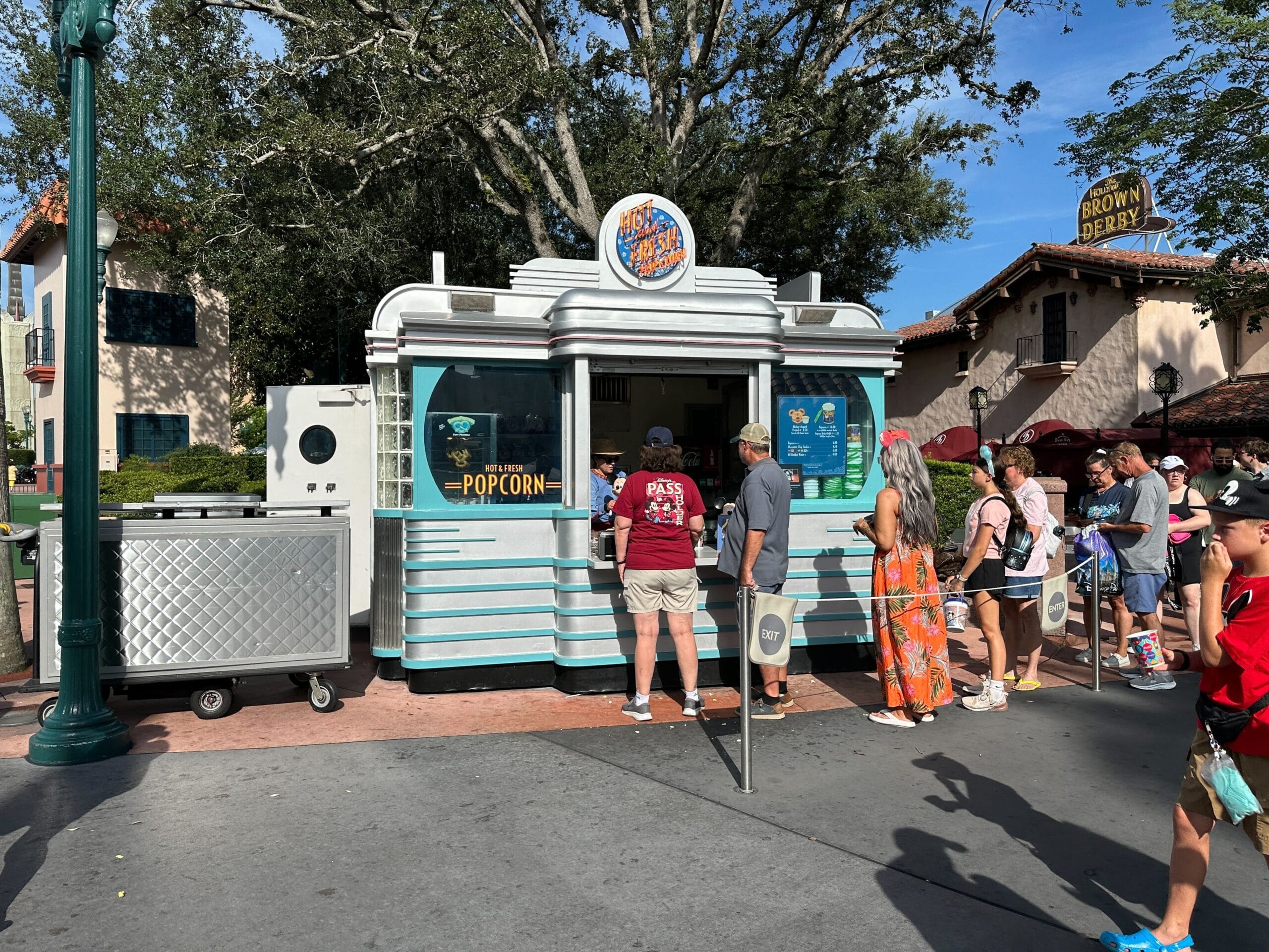 People wait in line at a retro-style popcorn food stand under sunny skies, surrounded by trees and a few buildings for talking mickey popcorn bucket