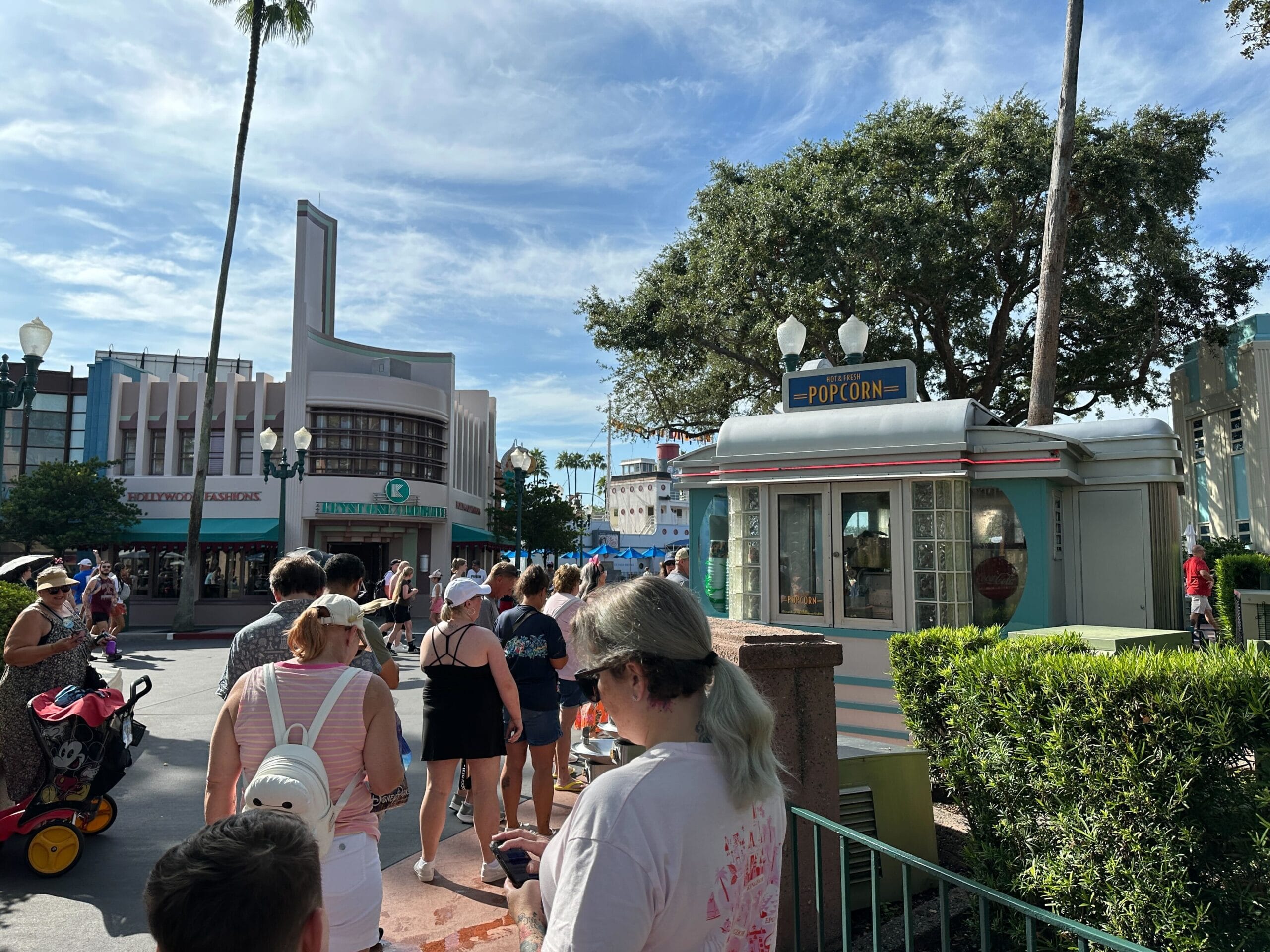 People wait in line at a popcorn stand in a sunny outdoor theme park with palm trees and art deco buildings.
