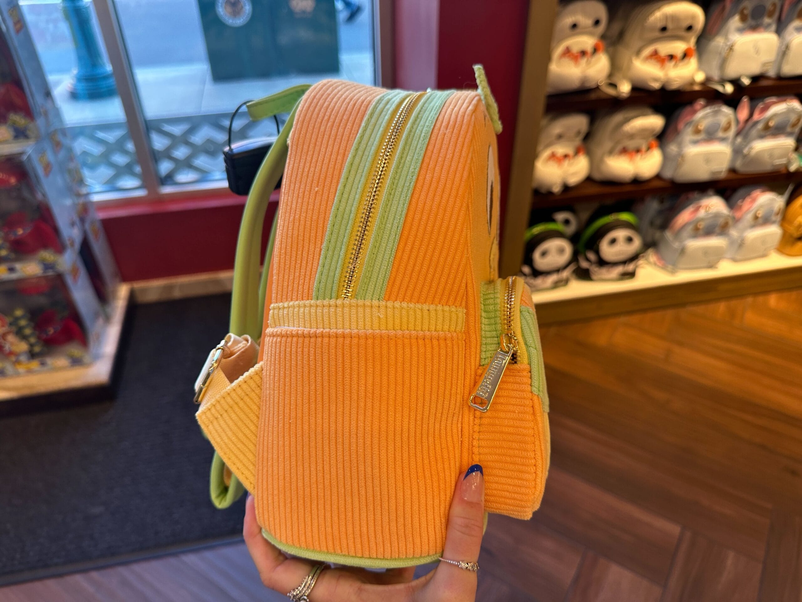 A hand holds an Orange Bird corduroy Loungefly backpack in a store, with plush character bags on shelves behind.