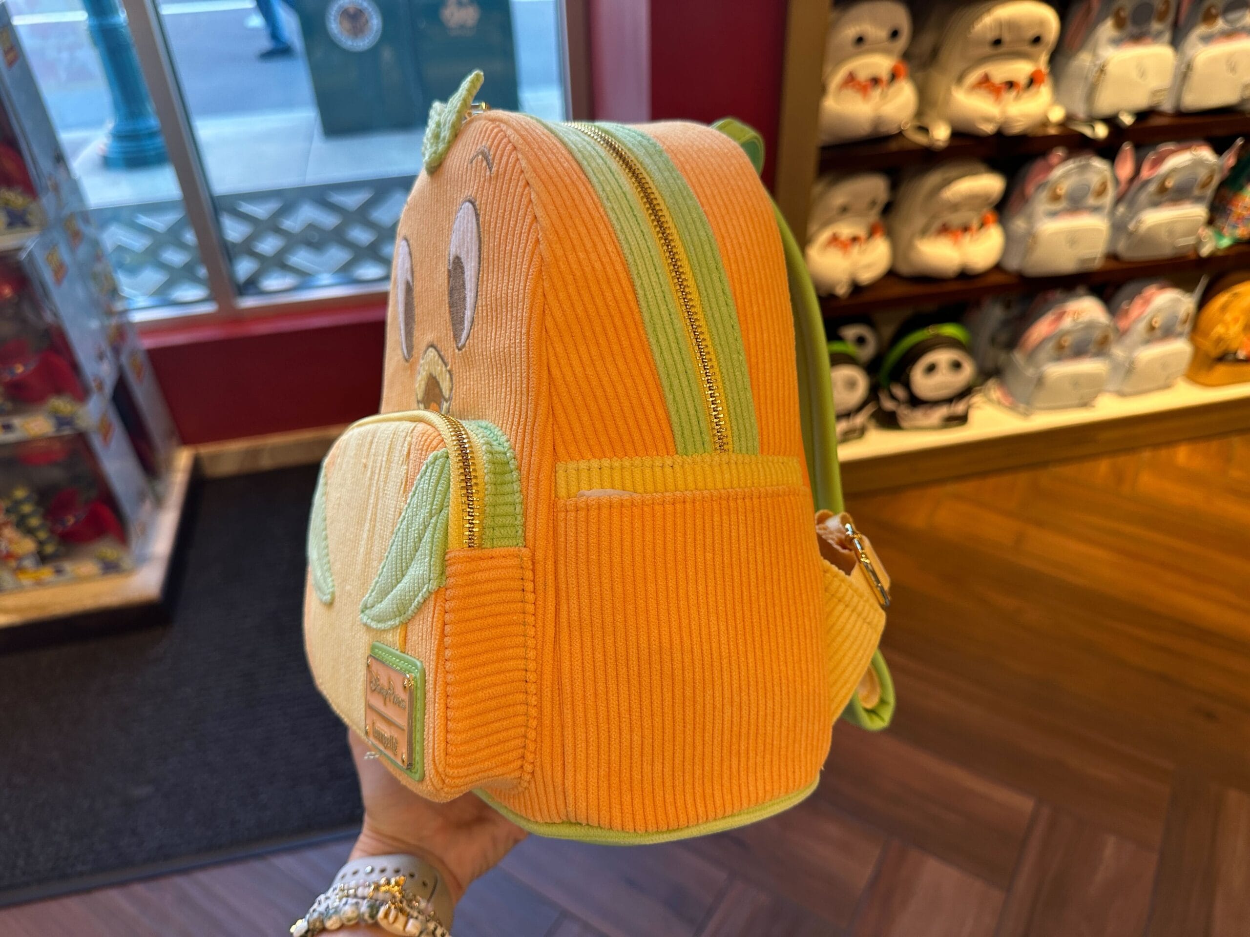 A hand holds an Orange Bird Loungefly backpack shaped like a cute character, surrounded by plush toys on store shelves.