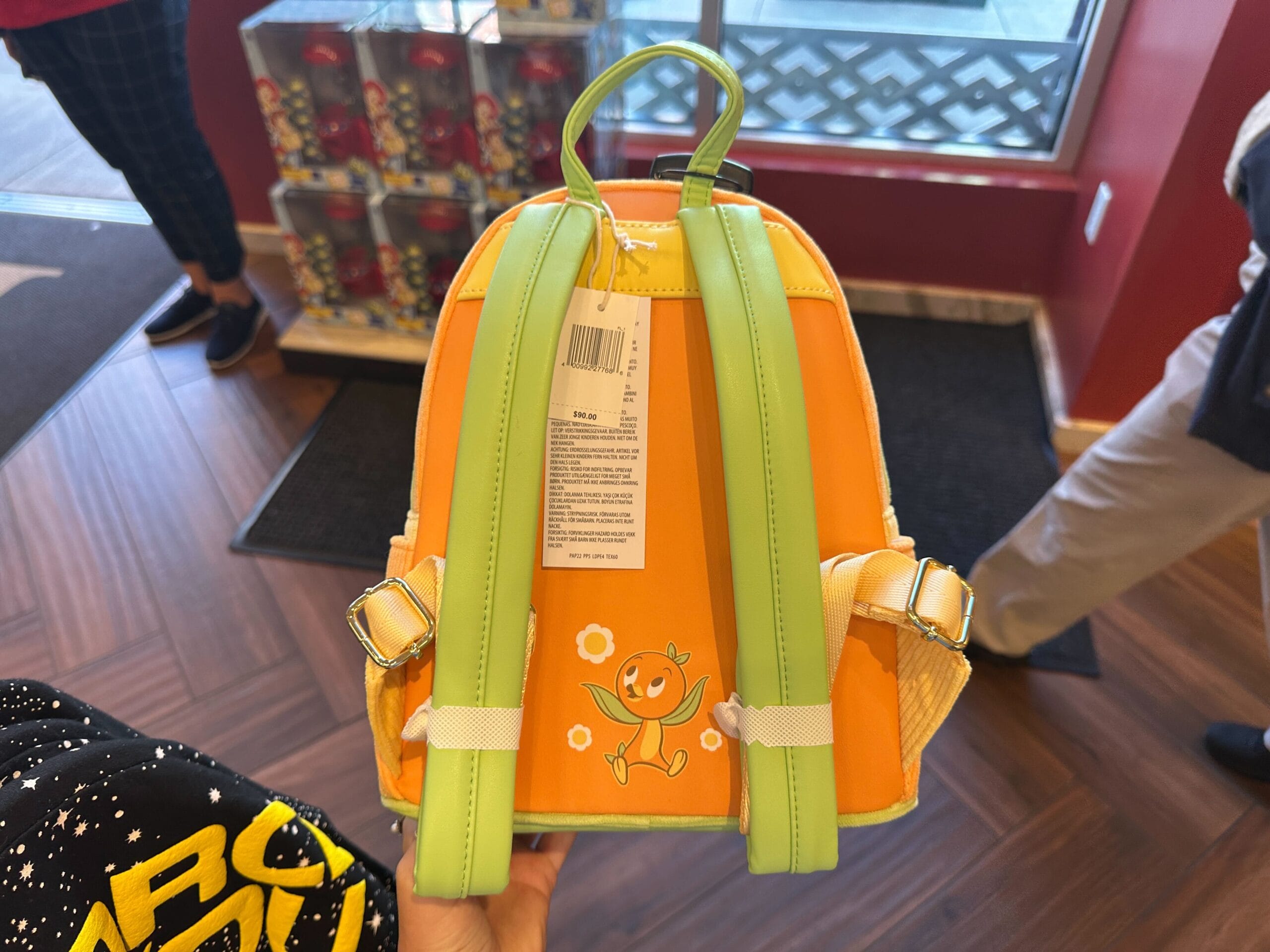 A small orange and green Loungefly backpack featuring a smiling Orange Bird cartoon, held in a store.