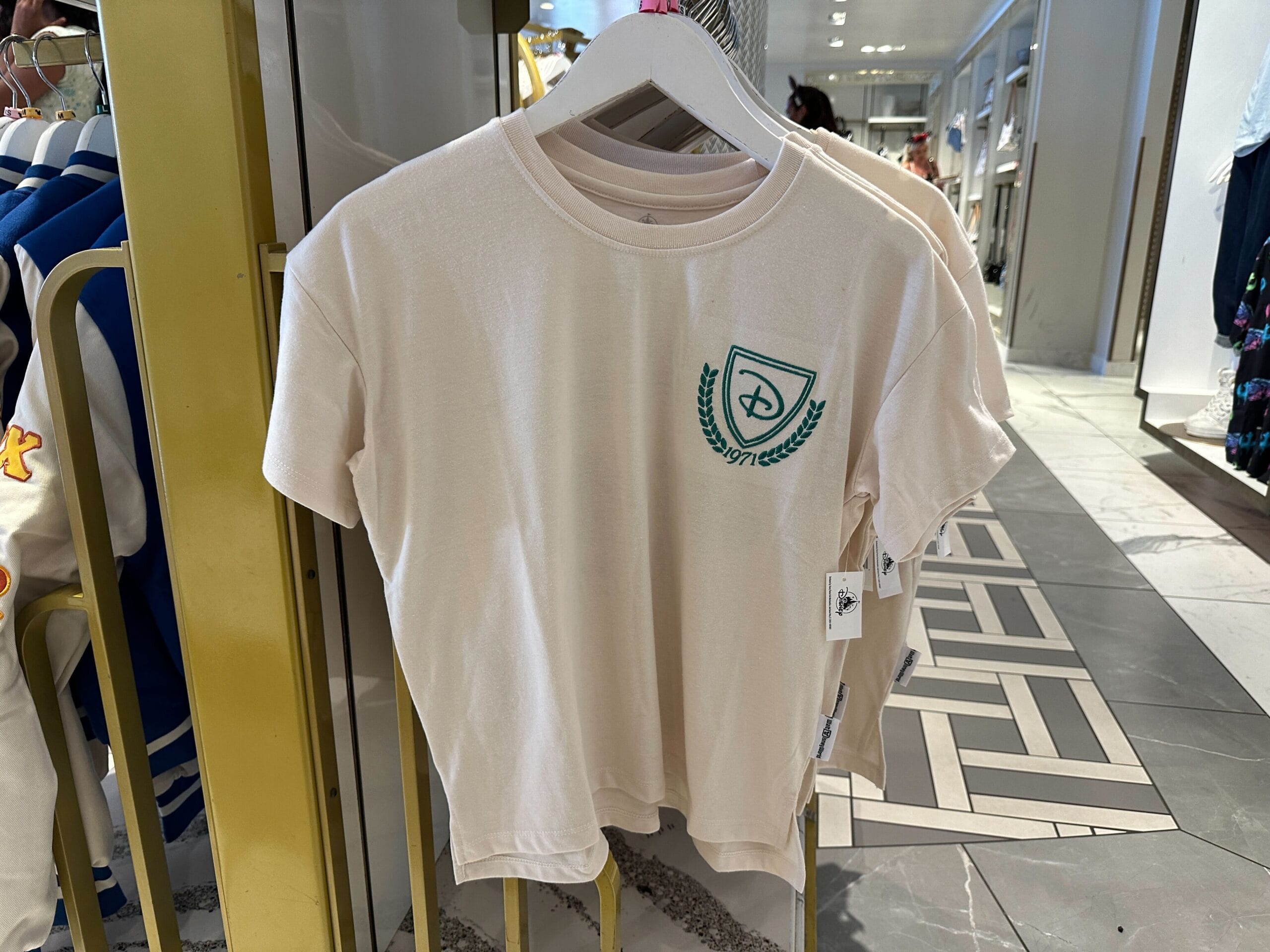 A white t-shirt with a green crest logo from Walt Disney World Apparel hangs on a rack in a clothing store.