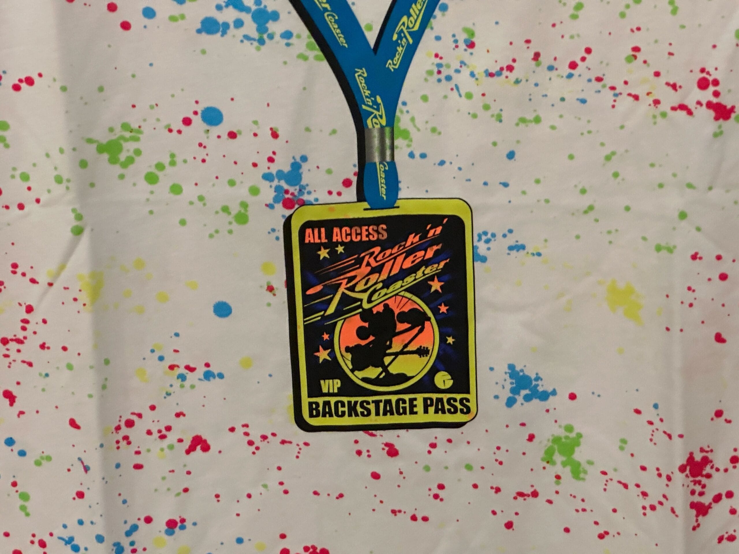 VIP backstage pass on a blue lanyard over a white shirt with colorful Rock 'n' Roller Coaster merch paint splatters.