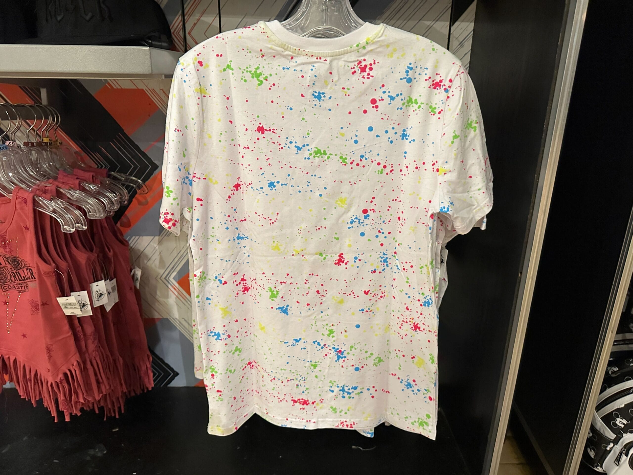 A white T-shirt with colorful paint splatters, inspired by Rock 'n' Roller Coaster, hangs on a store rack.