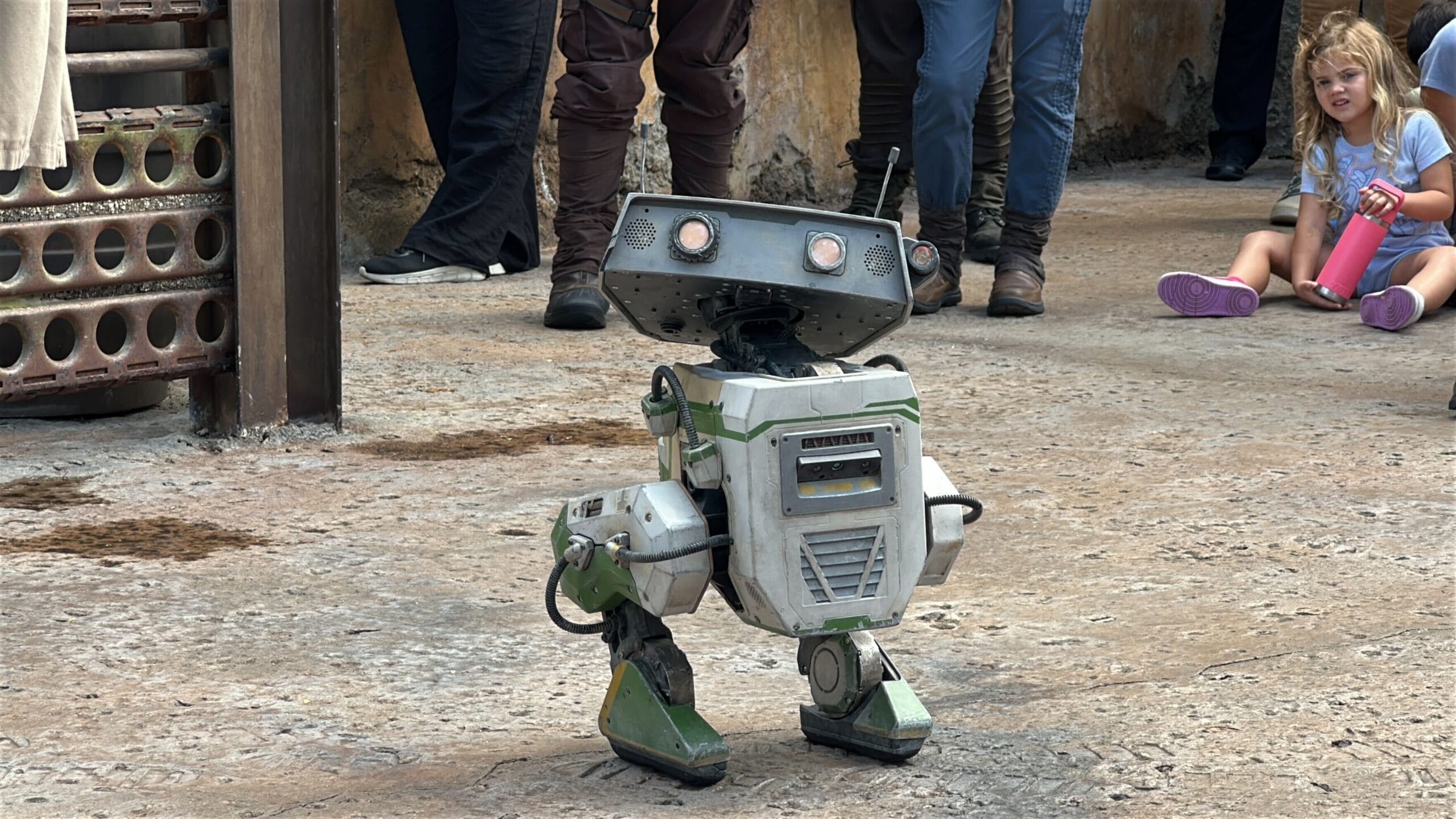 A small bdx droid with green and white panels stands on concrete at Hollywood Studios, people and a child nearby.