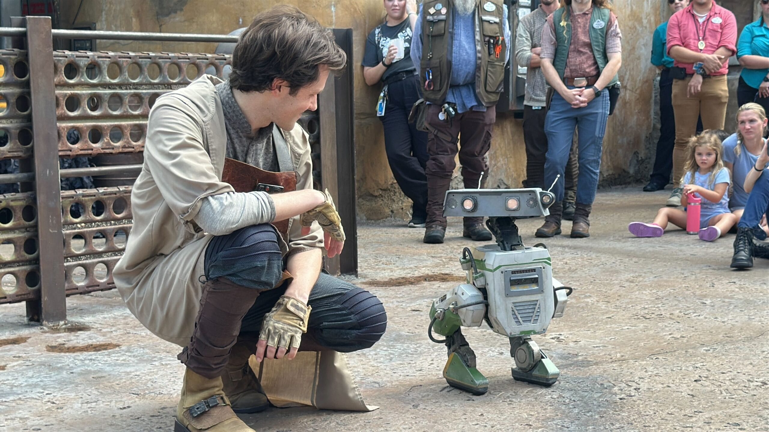 A man in costume kneels and smiles at a bdx droid, with people watching at Hollywood Studios in the background.