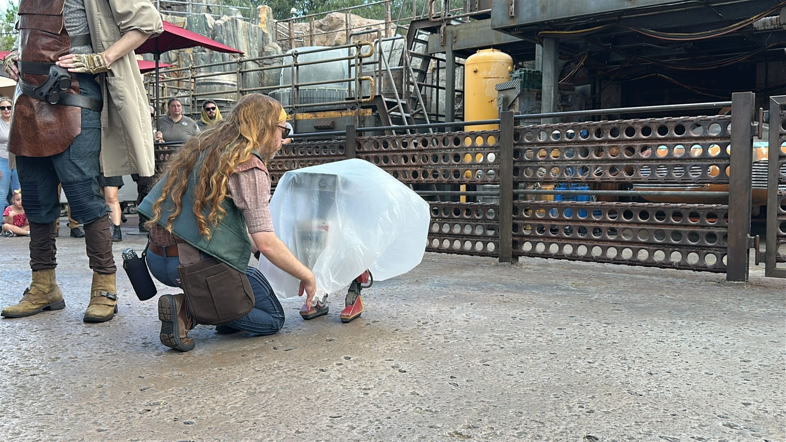 A woman crouches beside a bdx droid covered in plastic near industrial decor at Hollywood Studios.